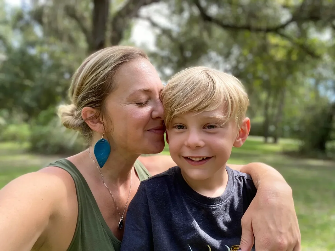 A woman with earrings and a necklace is hugging a smiling young boy with blonde hair outdoors, with trees and greenery in the background.