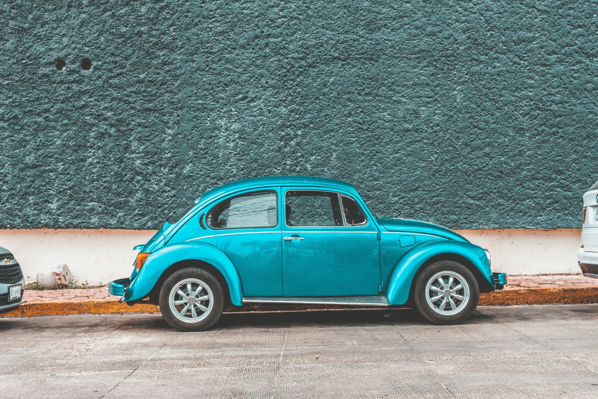 A vintage blue Volkswagen Beetle car parked on a city street against a textured teal wall.