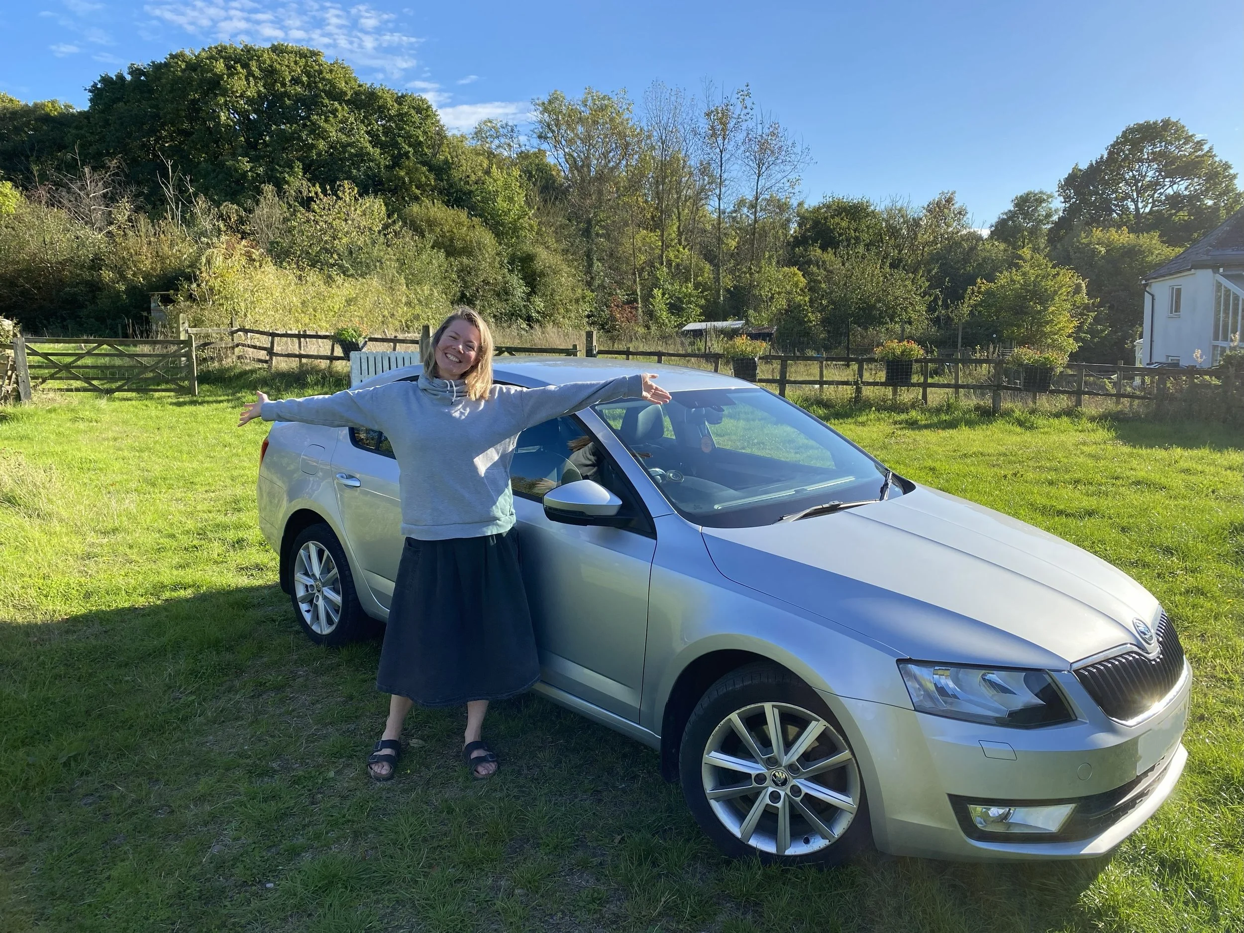 A woman with her arms outstretched standing next to a silver car in a grassy field with trees and a wooden fence in the background.