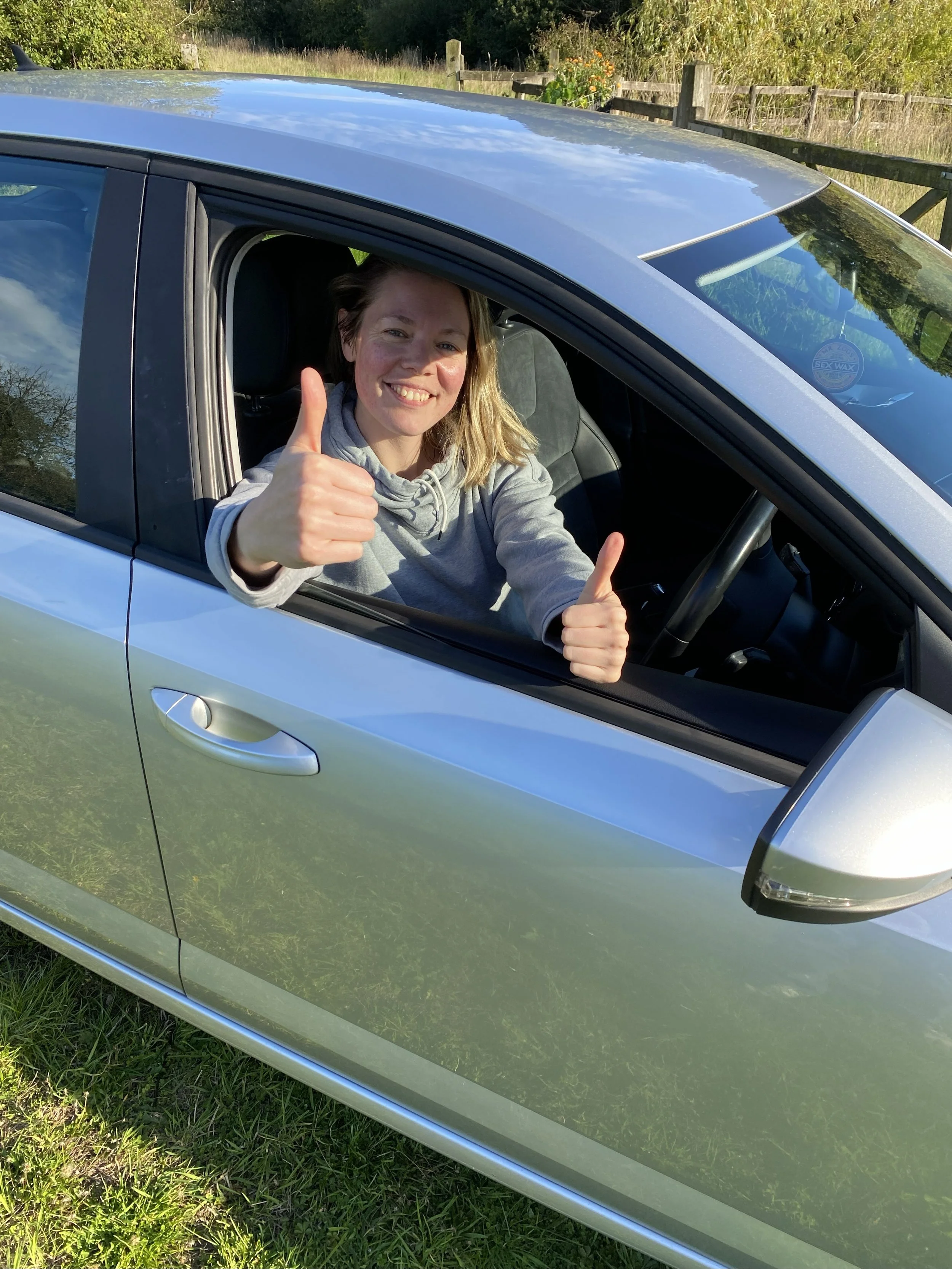 A woman sits in a silver car, smiling and giving two thumbs up through the window.