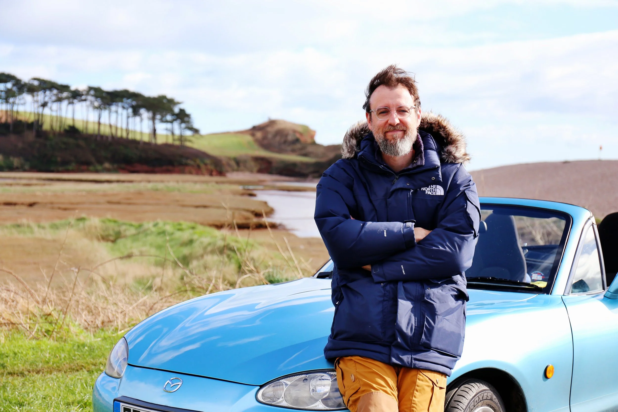 A man with glasses, a beard, and a mustache stands outdoors next to a blue convertible Mazda car with arms crossed, wearing a blue winter jacket with a fur-lined hood and mustard-colored pants. Behind him are grassy fields, some ponds, and a hill with trees under a partly cloudy sky.