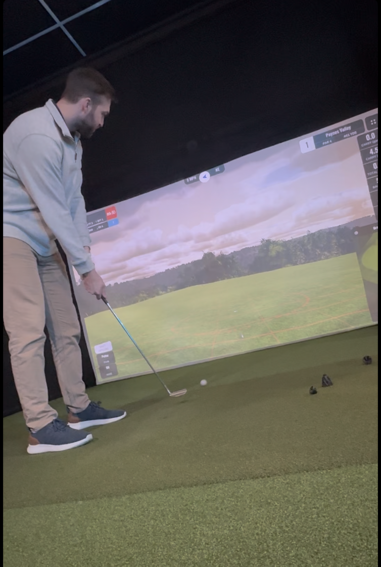 A man at an indoor golf simulator hitting a golf ball with a club, with a virtual golf course displayed behind him.