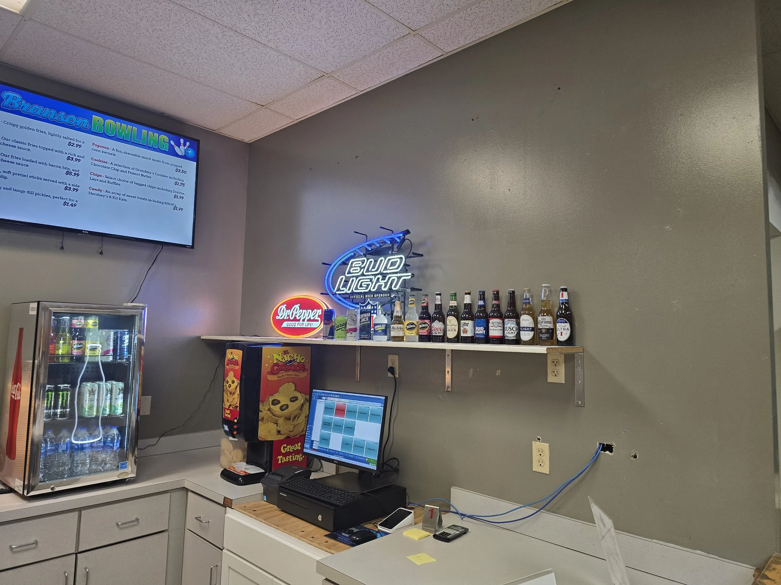 A bar area with a shelf holding various beer bottles, a neon sign that says 'Bud Light', a small fridge with drinks, a computer, a candy dispenser, and a mounted TV screen displaying a menu.