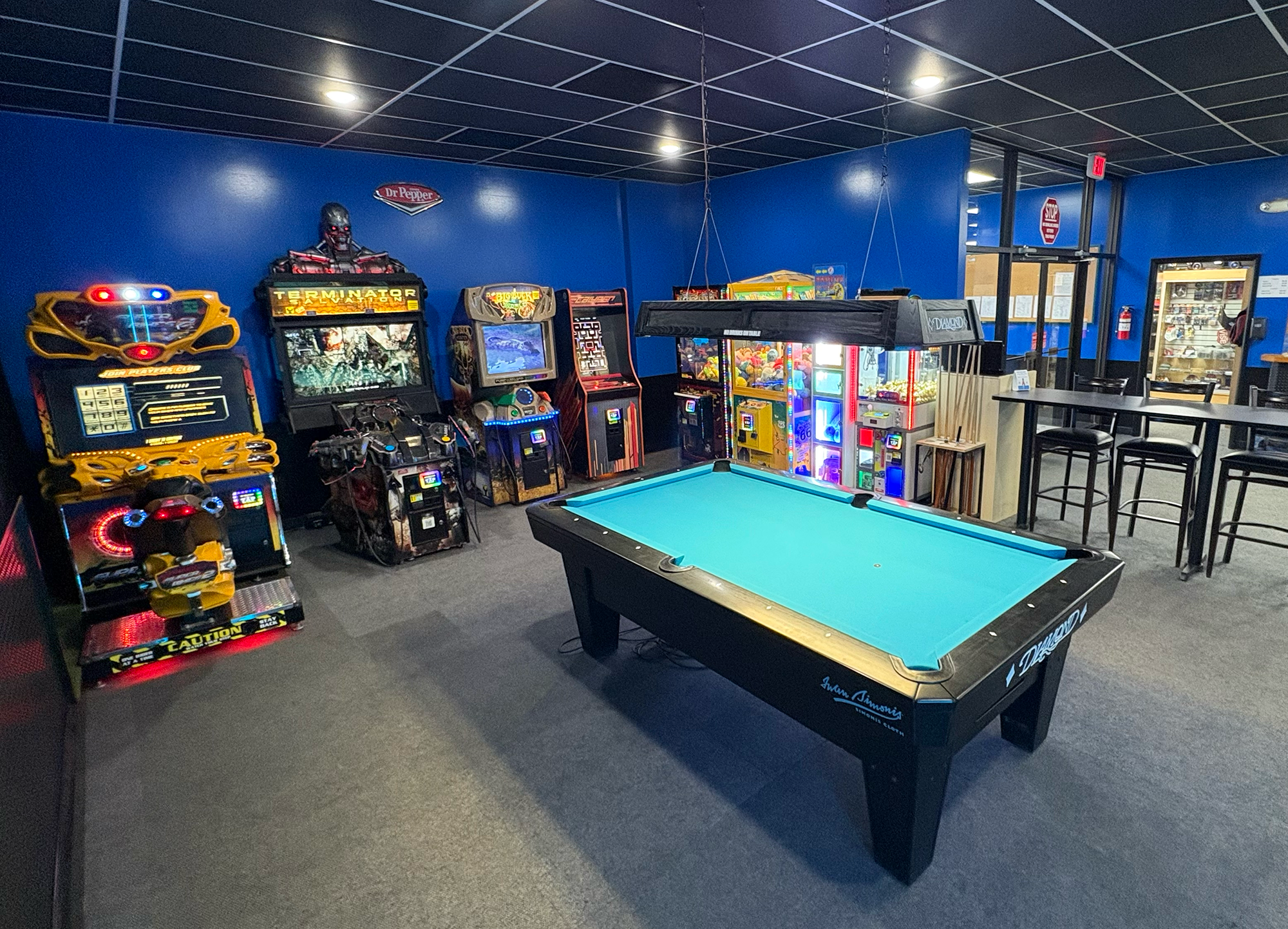 An arcade room featuring vintage arcade machines, a pool table, and consoles, with a blue wall and black ceiling.