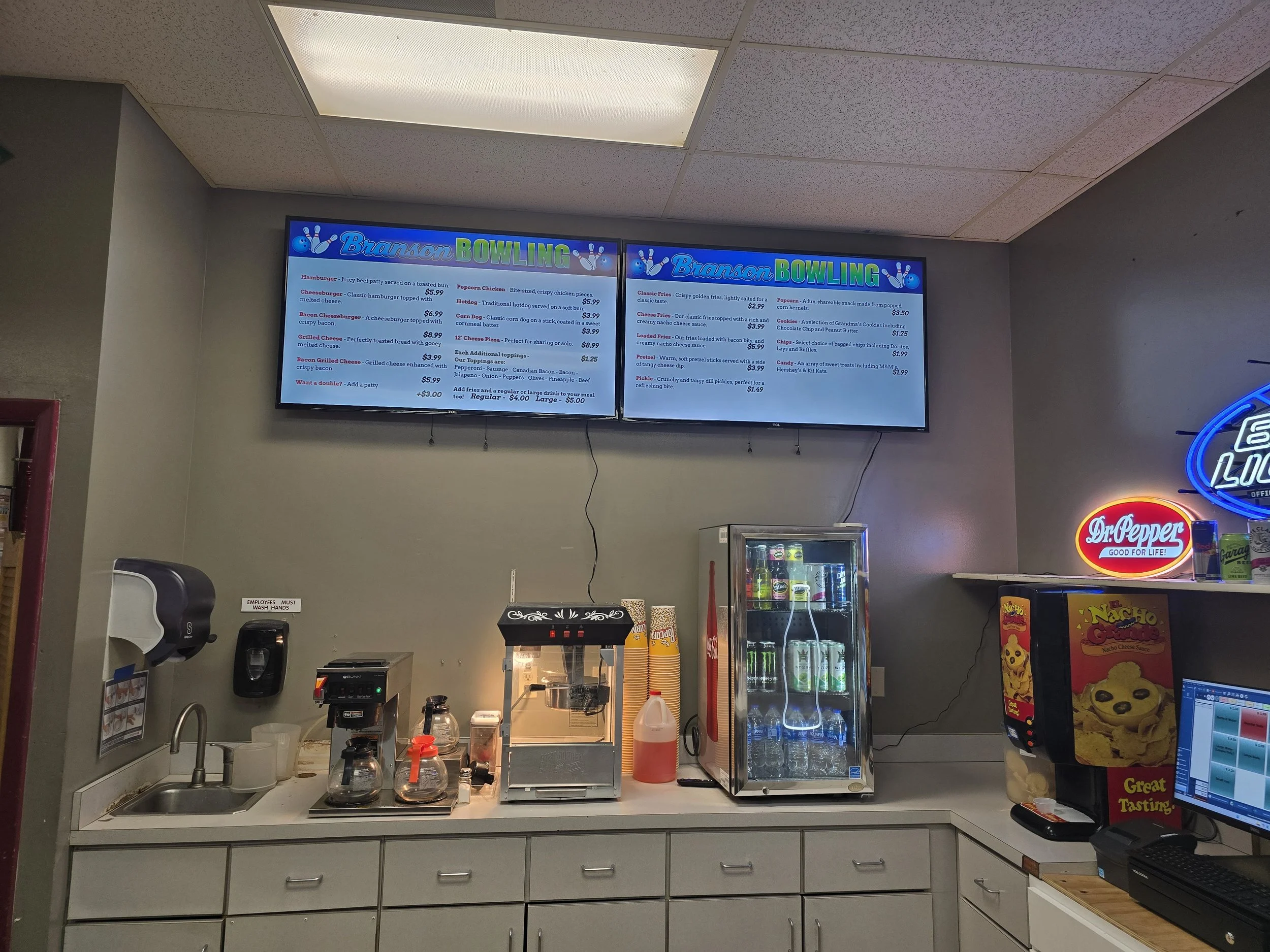 A counter at a bowling alley with a coffee machine, soft drink refrigerator, and a nacho cheese dispenser. Behind the counter, there are digital menu boards displaying food and drink options. To the right, neon signs for Dr. Pepper and other brands are visible, along with a computer monitor.