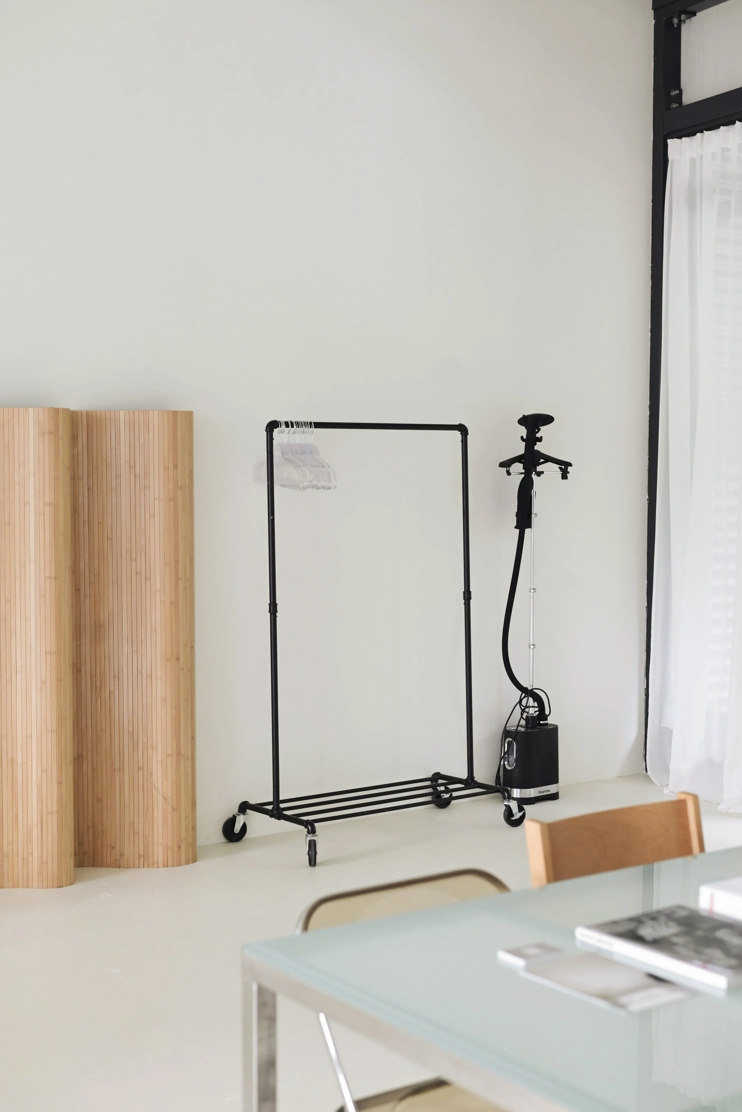 Empty clothing rack on wheels with a few clear hangers, bath steam device connected to a power source, and part of a dining table with books and a chair, in a bright room with white walls and curtains.