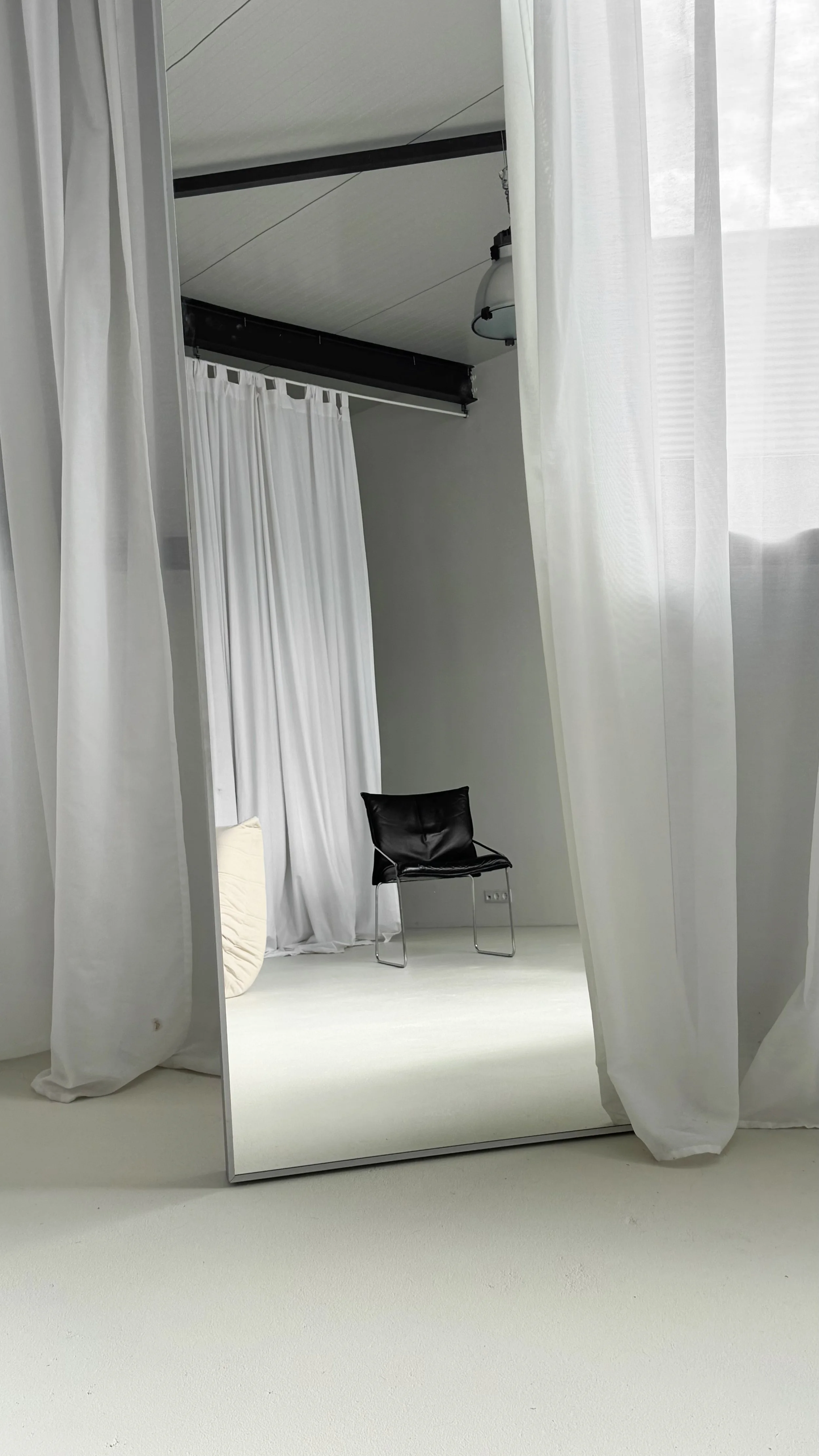 A black chair reflected in a full-length mirror with white curtains and a white wall in the background.