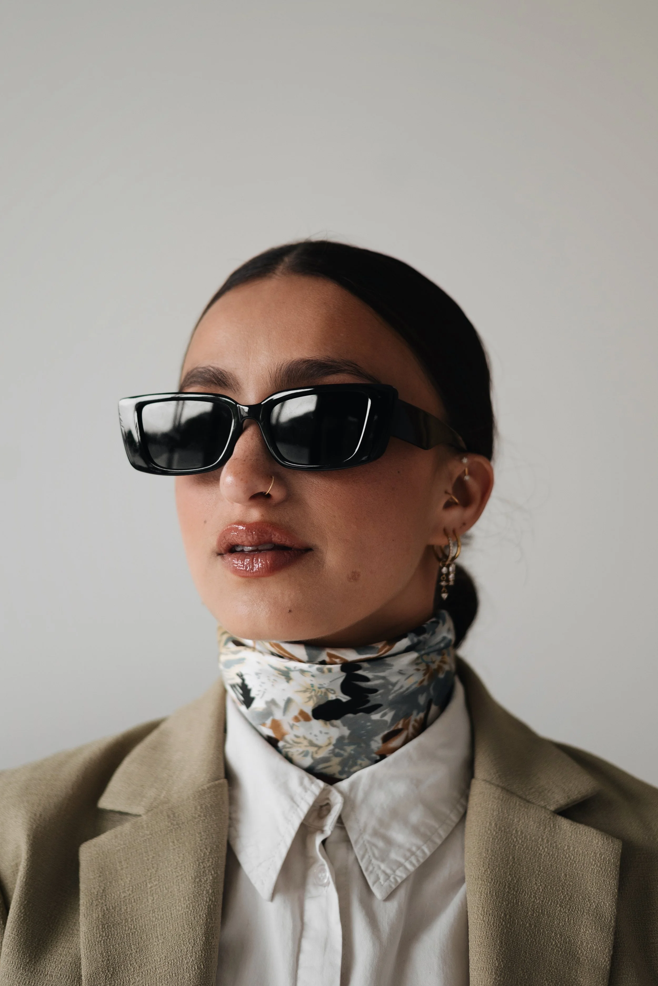 A woman wearing black sunglasses, gold earrings, a floral scarf, a beige blazer, and a white shirt, facing slightly to the left against a plain light background.