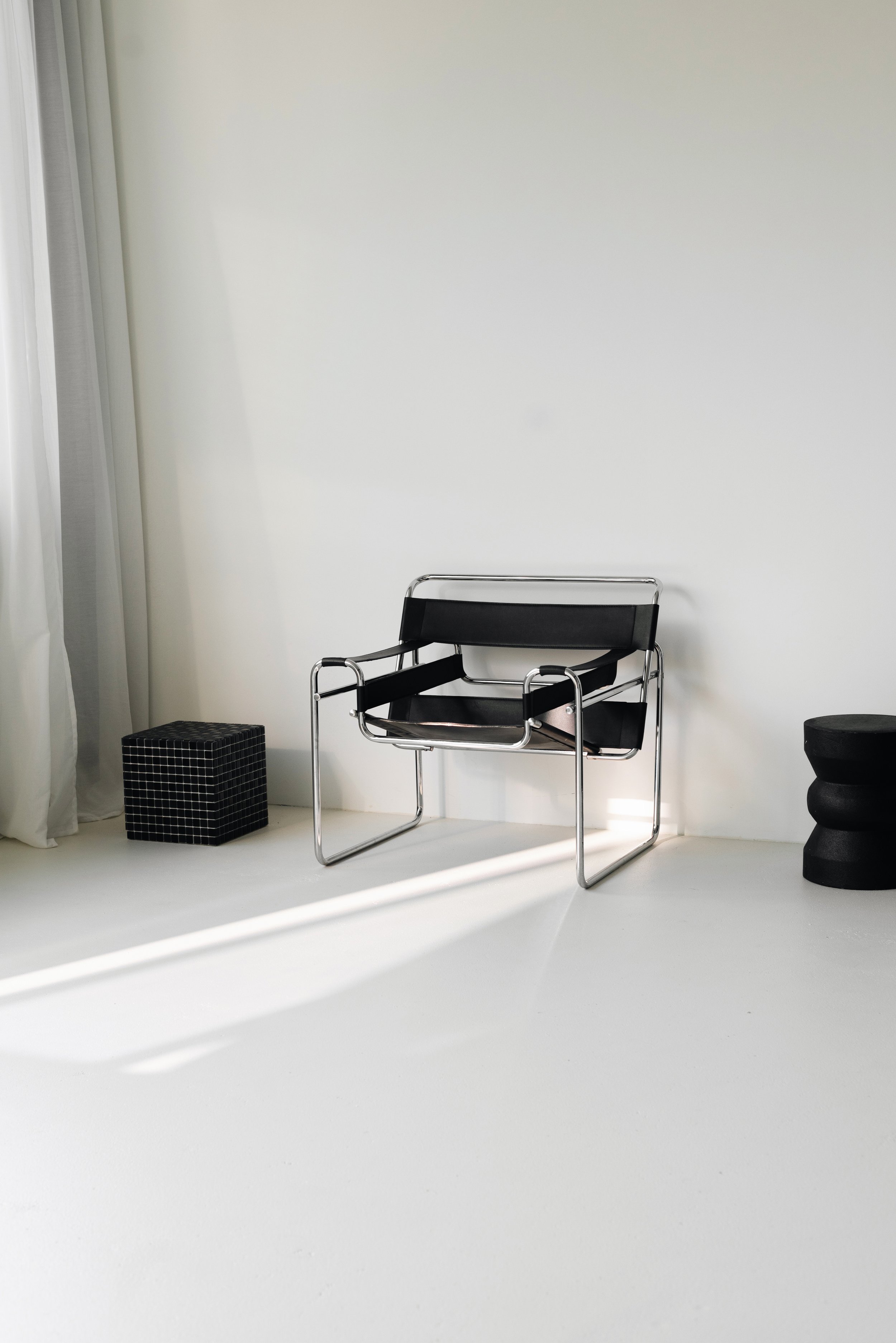 Minimalist interior with black chair, small black grid-patterned side table, and black sculptural stool near a white wall and curtain, illuminated by sunlight.