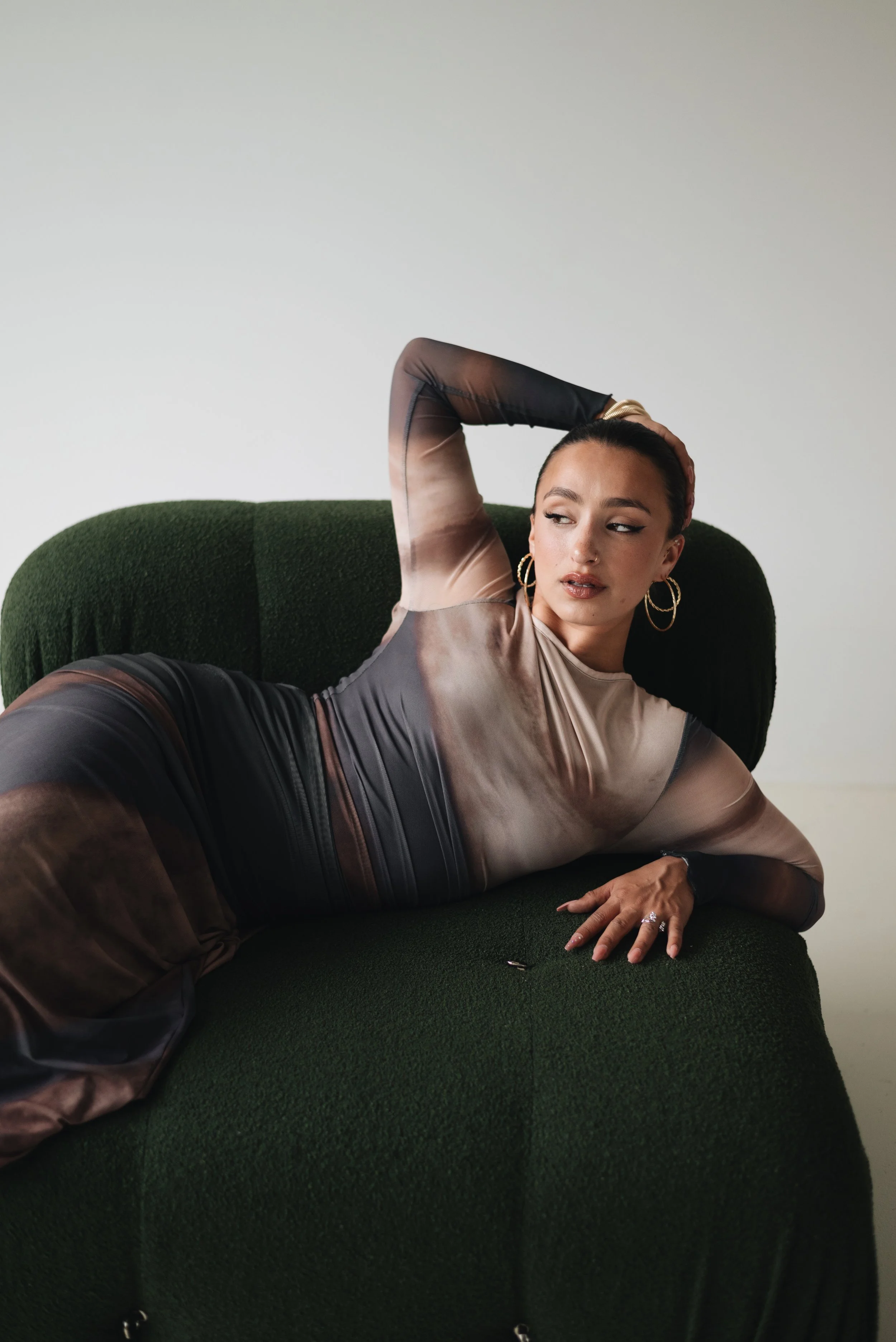 A woman lying on a dark green textured couch, resting her head on her left hand, wearing a sheer, long-sleeved, multicolored dress with shades of beige, brown, and black, and large hoop earrings, with a neutral expression against a plain white wall b