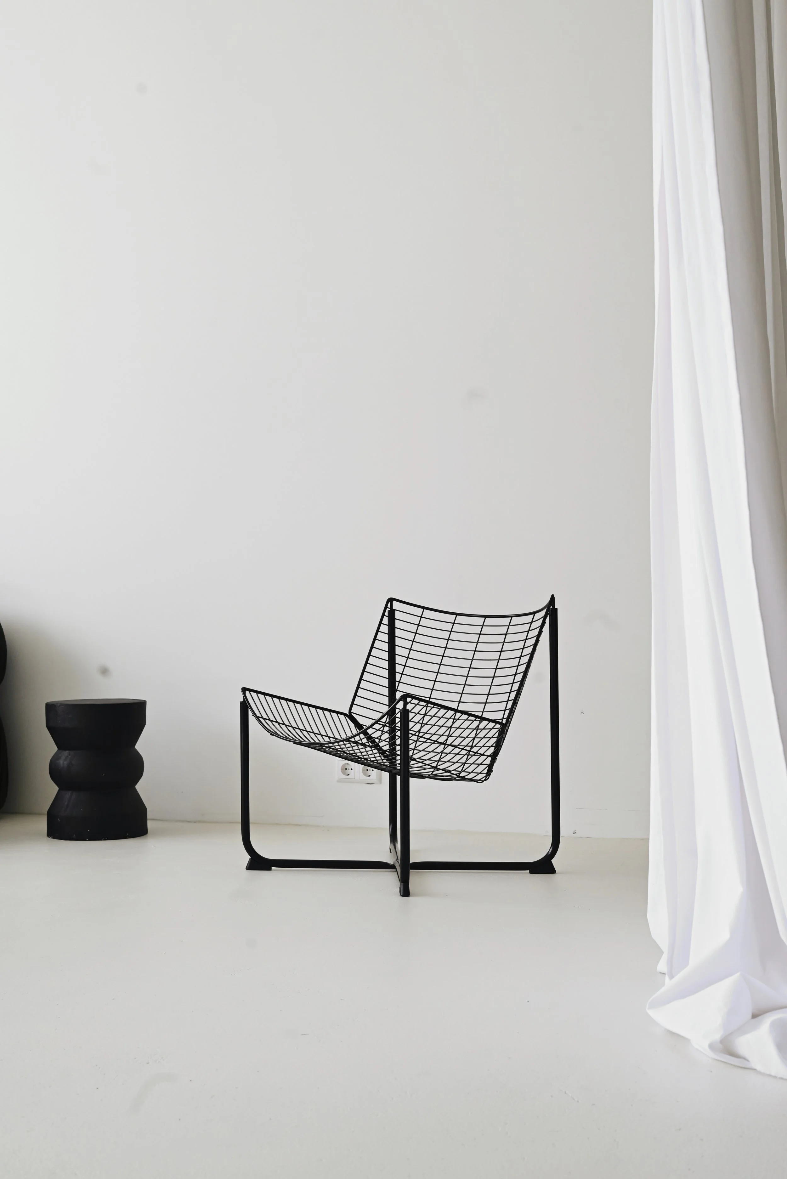 A black wireframe chair next to a black abstract side table, set against a plain white wall with a plug socket, and white curtains on the right side.