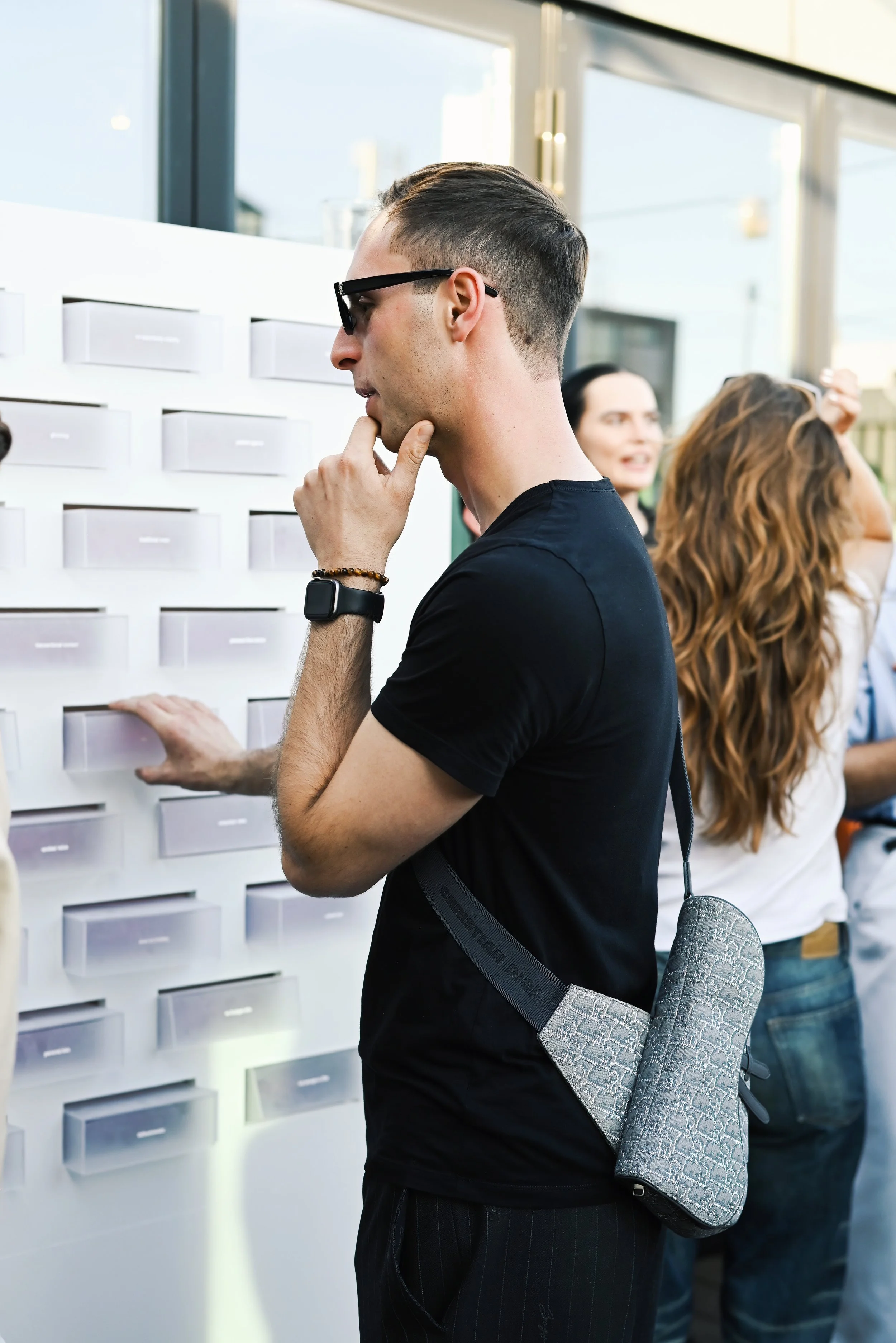 A young man wearing glasses, a black t-shirt, and a smartwatch is looking at a wall-mounted display with small white compartments, touching his chin with his hand in a thoughtful pose. Several other people are visible in the background.