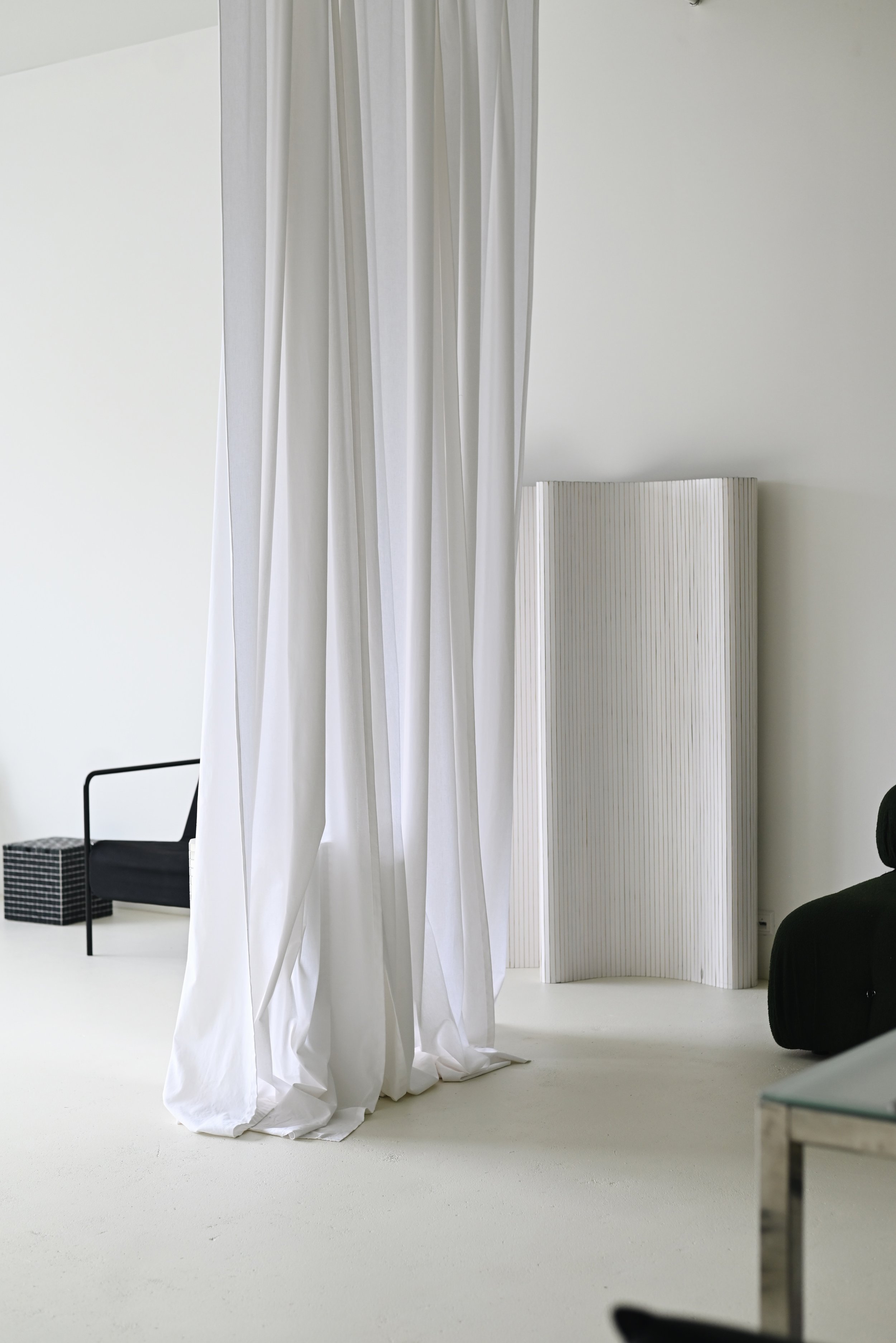 Minimalist interior with white walls, white curtain, black chair, wicker basket, white folding screen, and black armchair.