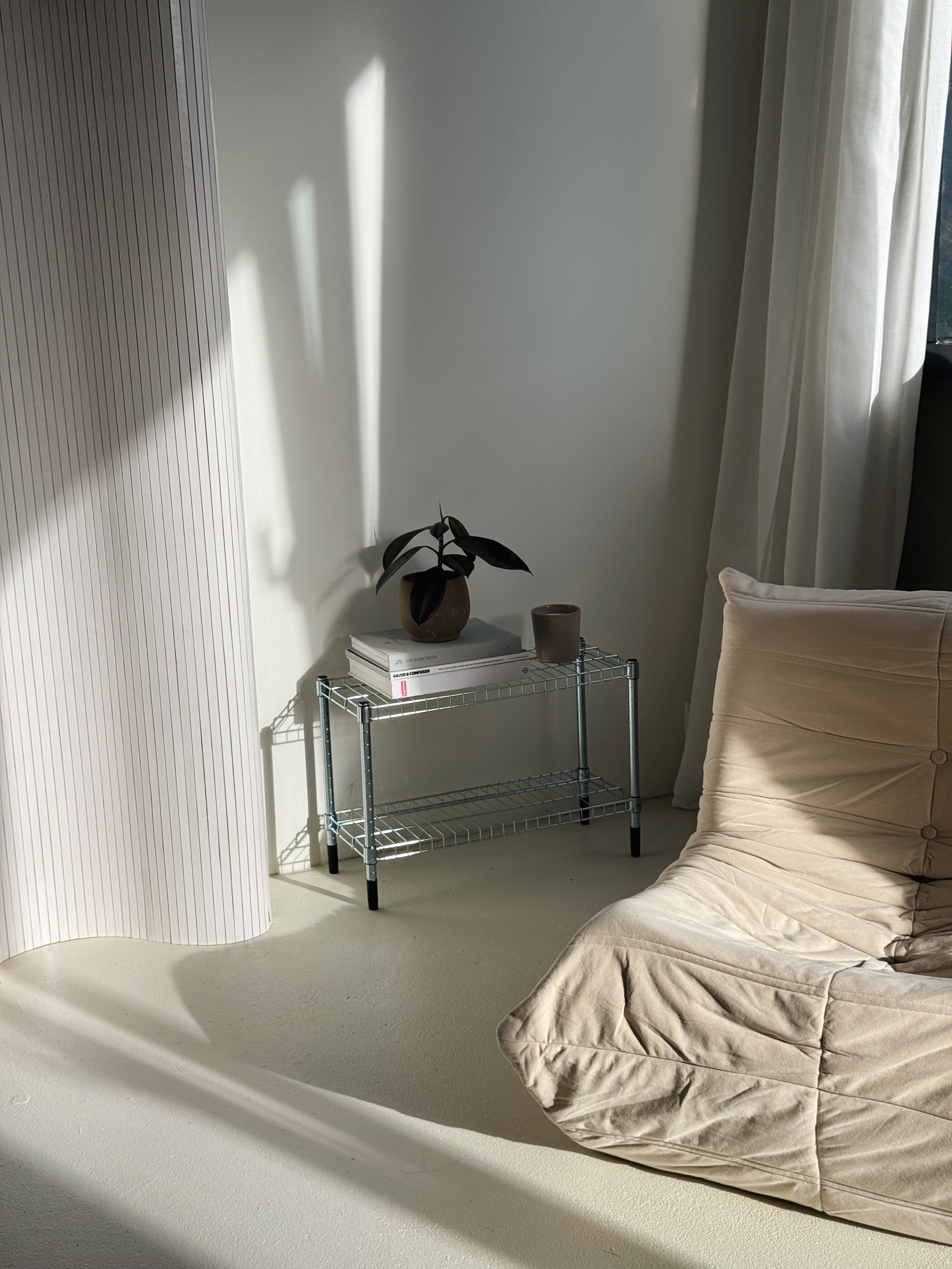 A corner of a room with a beige sofa, a small metal table with a potted plant, books, and a candle, and a window with white curtains letting in sunlight.