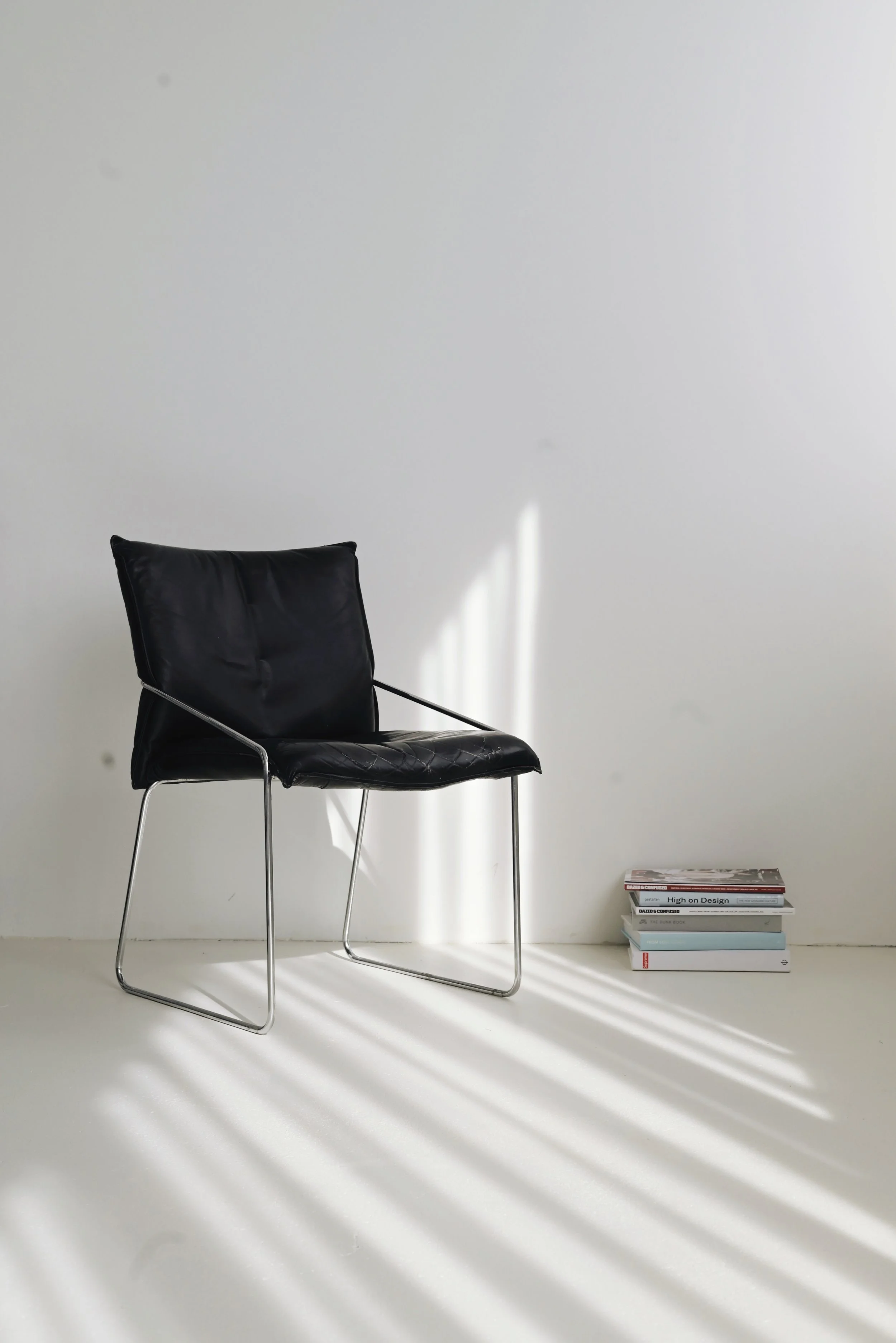 A black leather chair with a metal frame next to a small stack of books on the floor, with sunlight creating shadows on a white wall and floor.