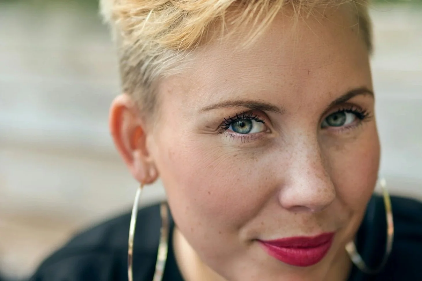 Close-up of a woman with short blonde hair, blue eyes, and red lipstick, wearing large gold hoop earrings.
