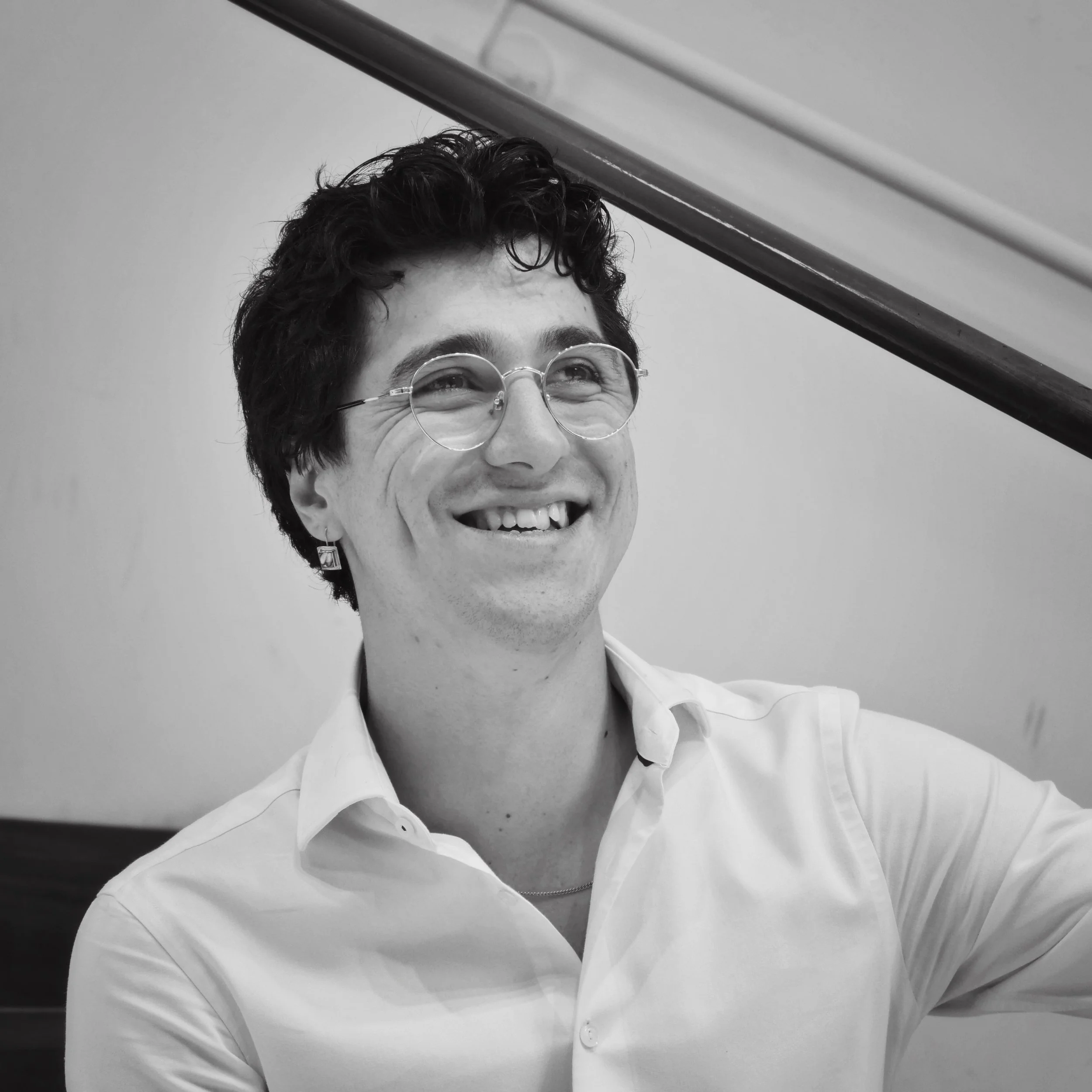 A man with curly dark hair, glasses, and earrings smiling cheerfully, wearing a white shirt, standing near a staircase.