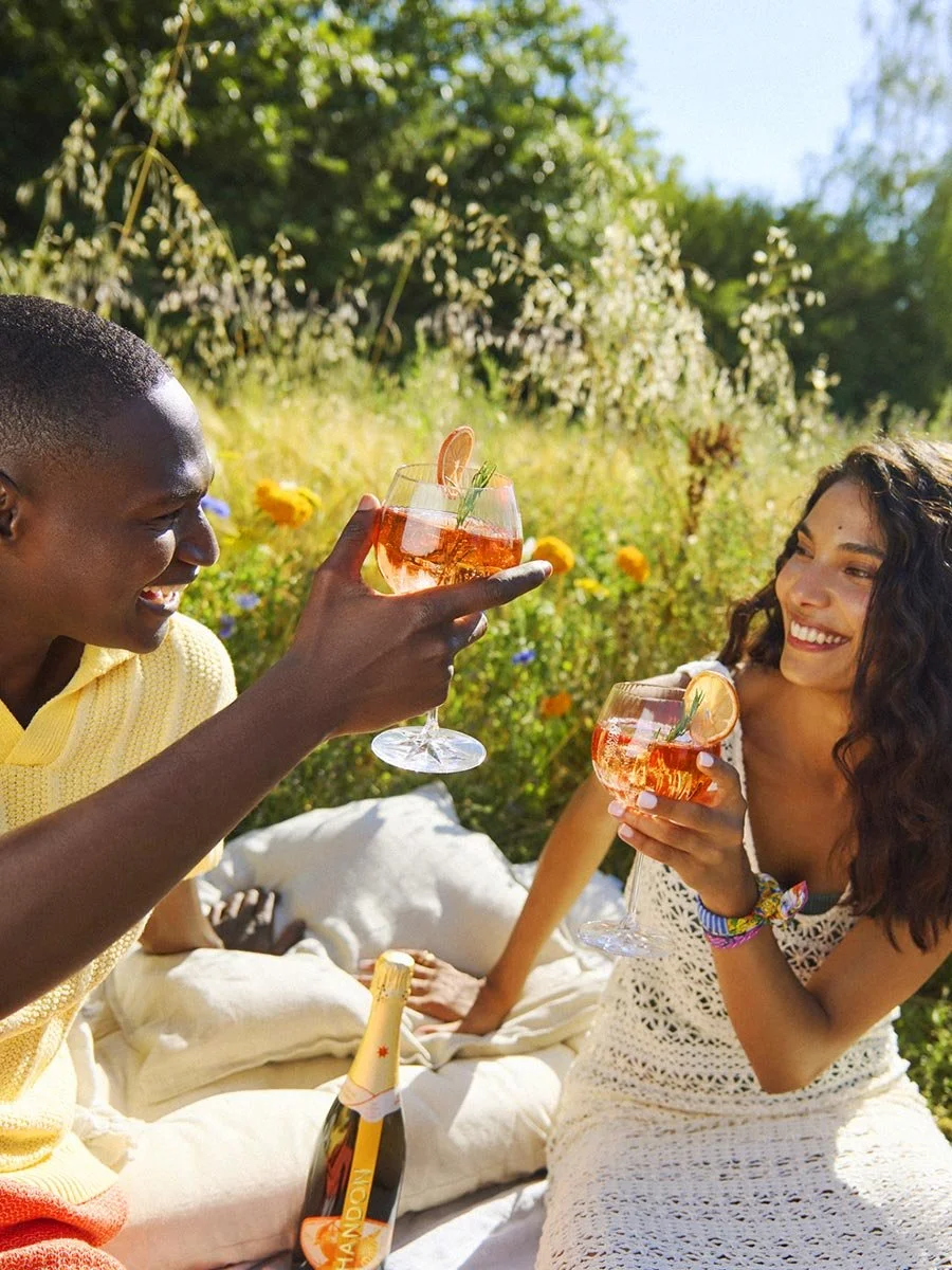 Deux personnes souriantes qui trinquent avec des verres à cocktail dans un extérieur ensoleillé, avec des fleurs et des arbres en arrière-plan. Campagne de l'agence de création L'ASSOCIÉ pour CHANDON.