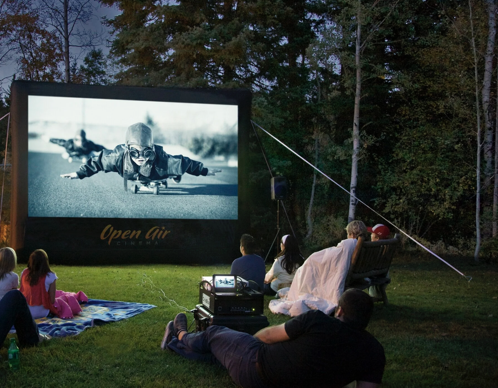 People watching an outdoor movie in a park at night on a large screen showing a child with goggles riding a skateboard.