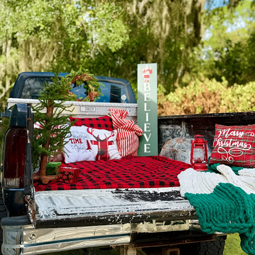 Decorated truck bed with Christmas pillows, a small Christmas tree, a lantern, a 'BELIEVE' sign, a 'Merry Christmas' pillow, and a green and white blanket, set outdoors with trees in the background.