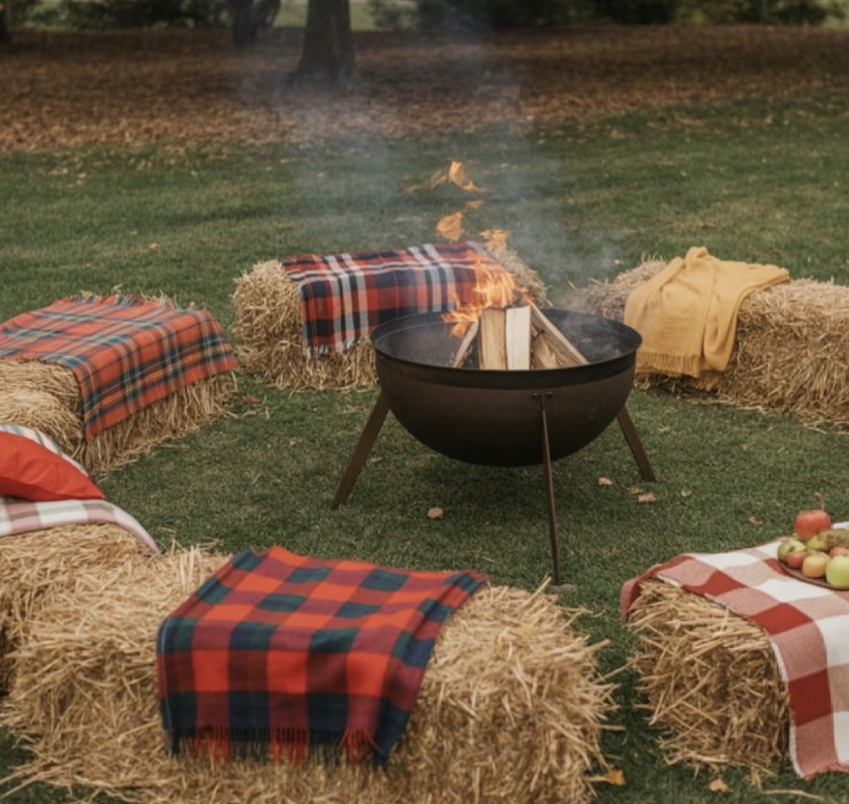 Hay bales with plaid and yellow blankets arranged in a circle around a barbecue fire pit on a grassy field, with apples and a small pumpkin on a table to the side.