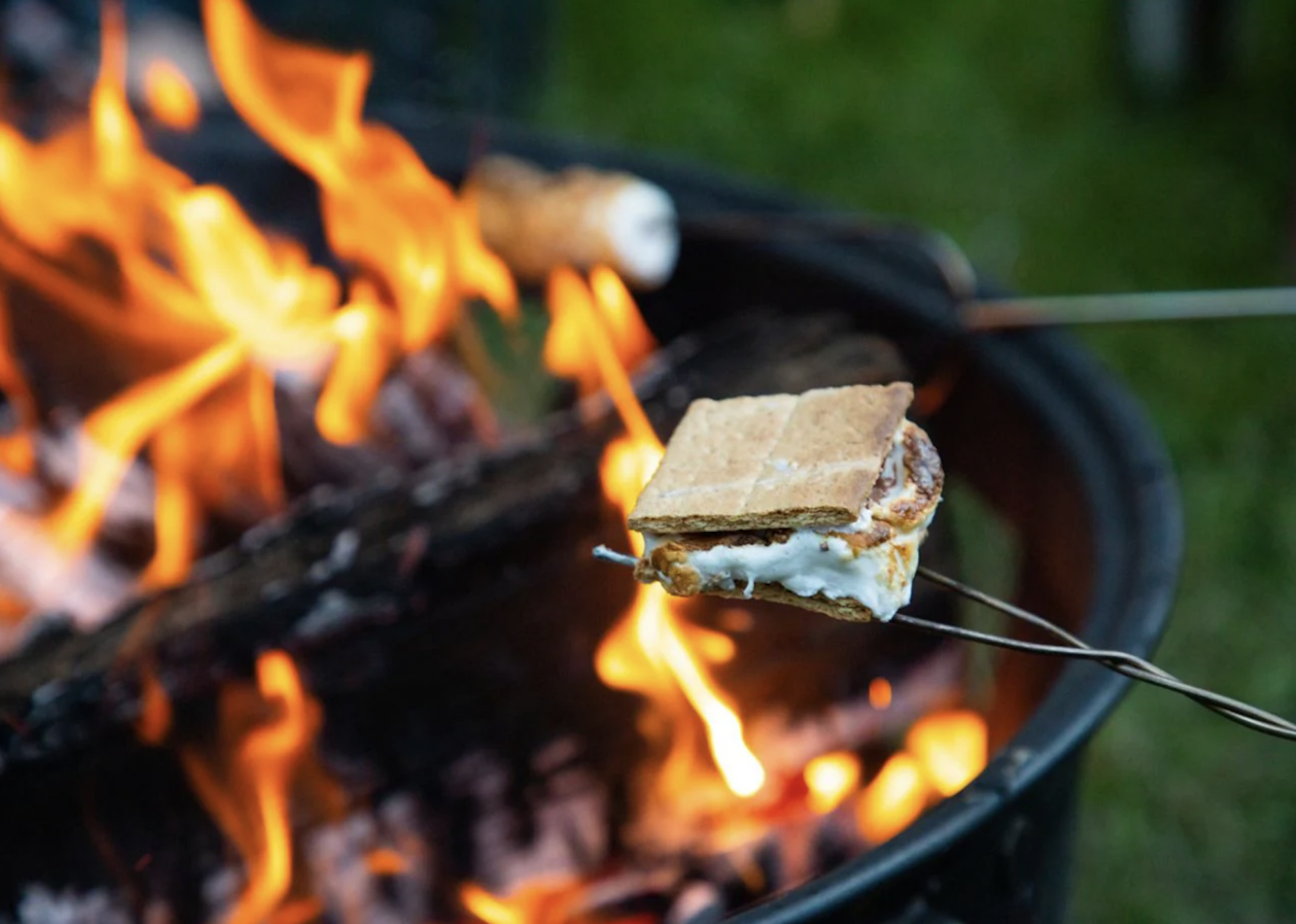A close-up of a s'more being roasted over an open fire, showing graham cracker, marshmallow, and chocolate with flames in the background.