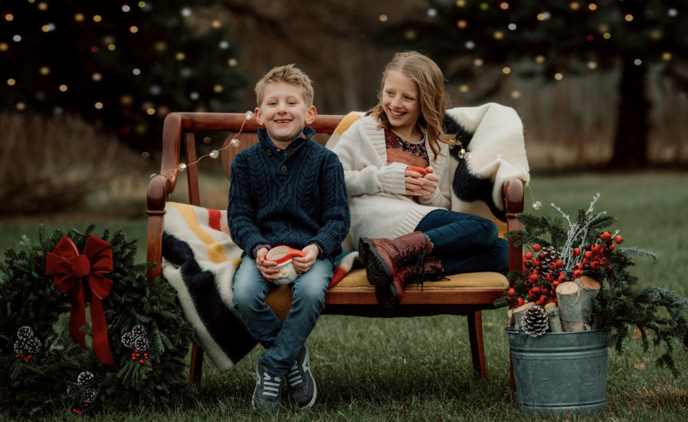 A boy and a girl sitting on a bench outdoors decorated for Christmas, holding mugs, with holiday wreaths, red berries, pine cones, and logs nearby.