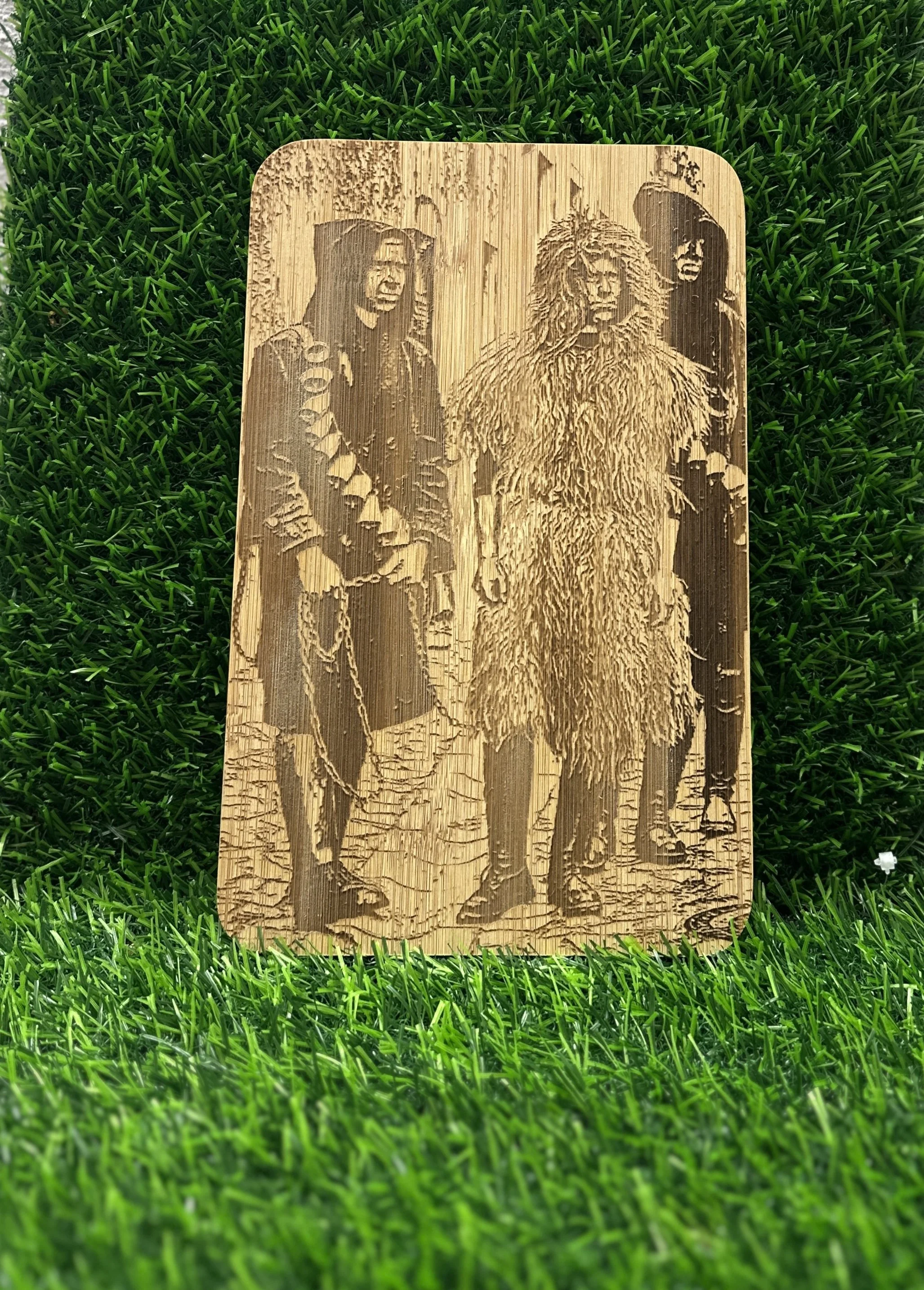 Insegna decorativa di legno inciso con tre figure umane tradizionali africane, posizionata su una superficie di erba verde.