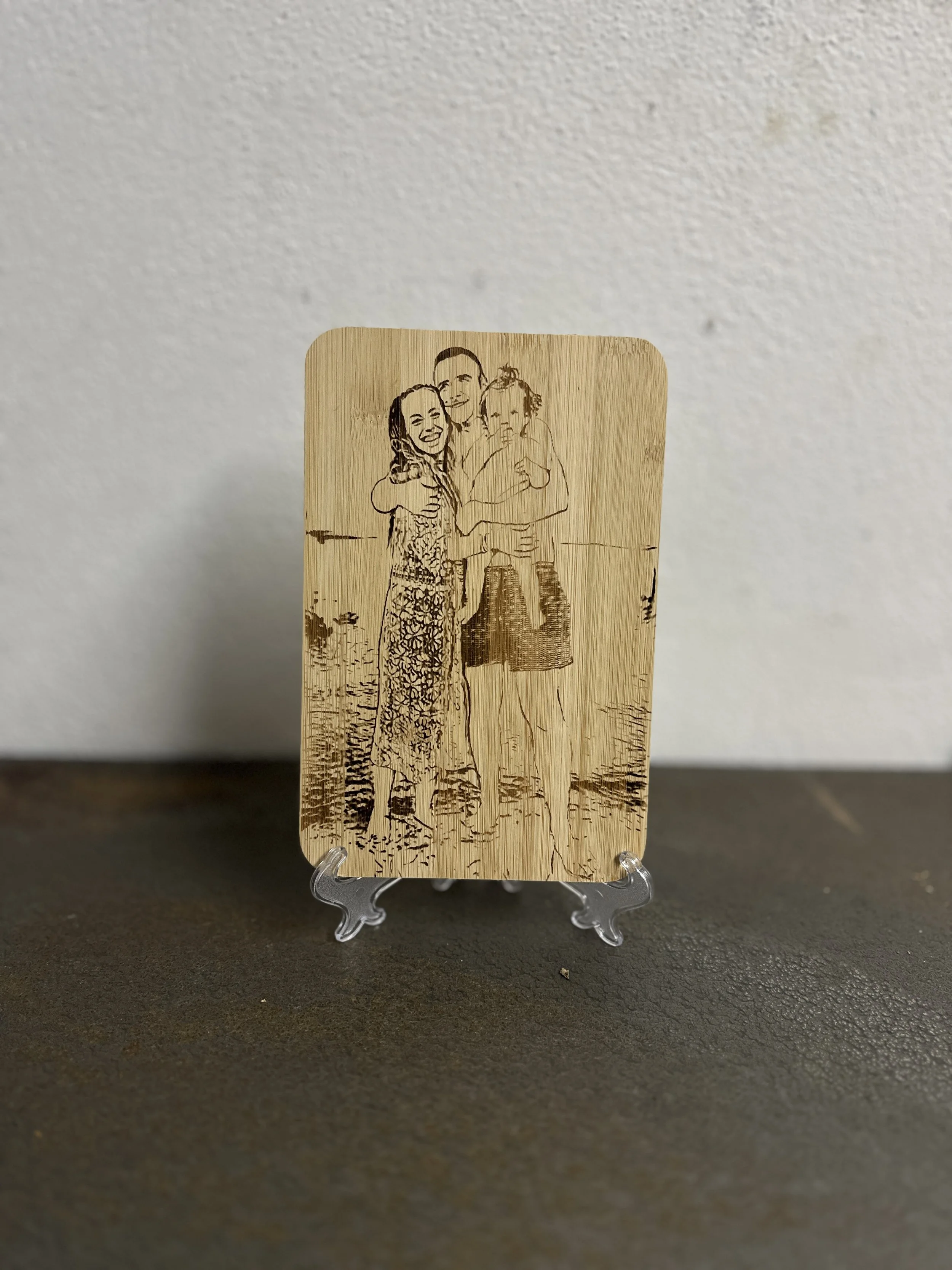 Segnalibro di legno con incisione di una famiglia che sorride e si abbraccia, posizionato su una superficie scura di fronte a una parete bianca.