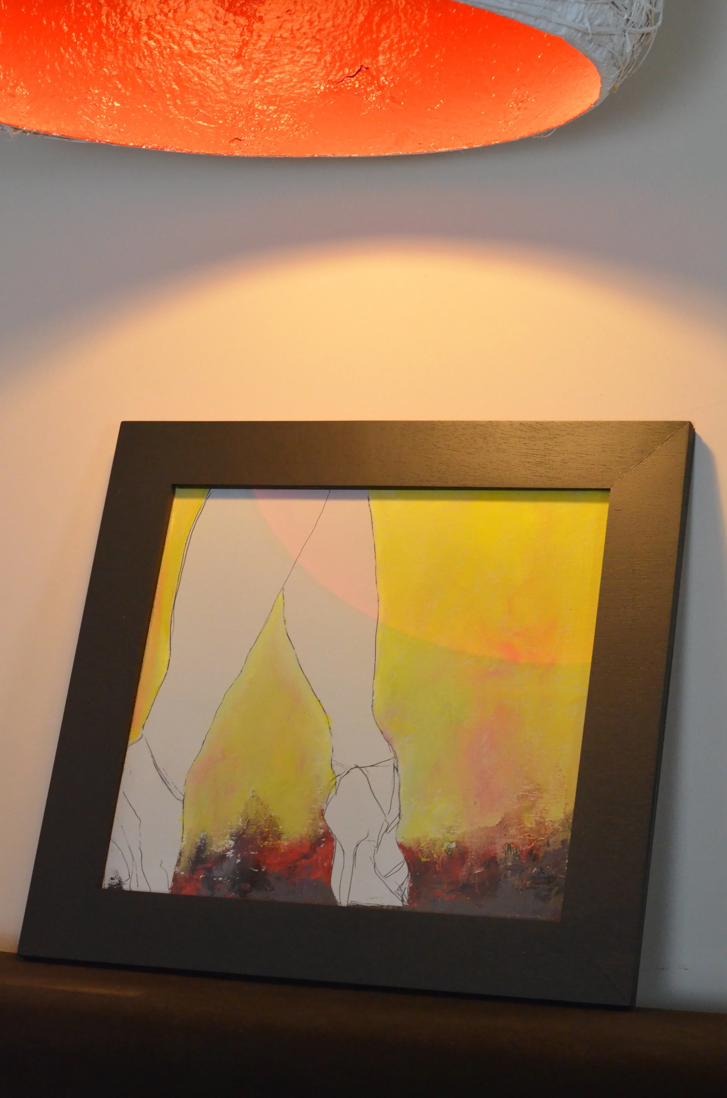 Quadro da delicadeza e leveza feminina com moldura escura, com uma pintura abstrata em tons de amarelo, vermelho e preto, com iluminação de uma luminária Polpa Lamps, quadro da série Humans, la fame qui danse. Autor LU.S