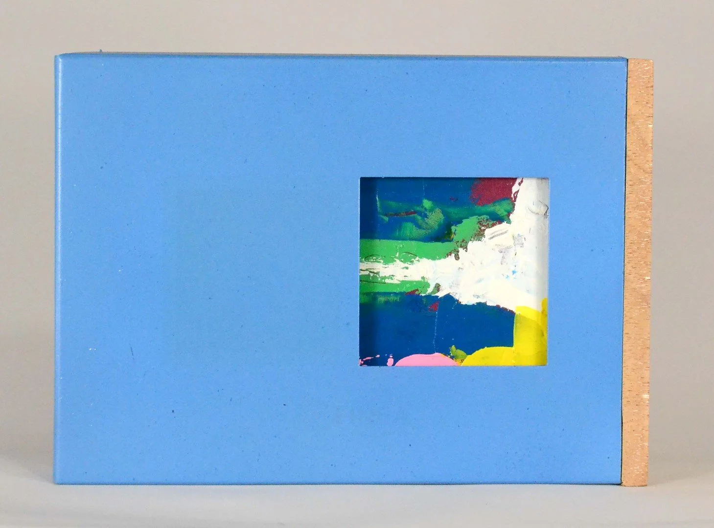 Caixa azul com uma janela pequena que revela uma pintura abstrata com cores verde, branco, azul, rosa e amarelo. Autor LU.S