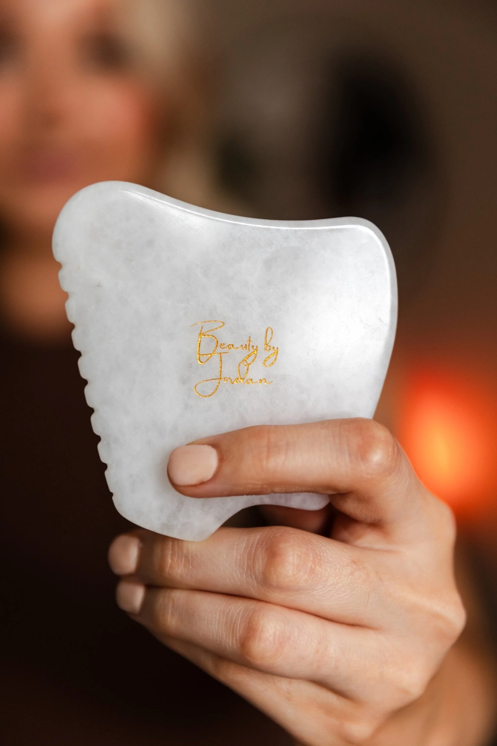 A person holding a white jade facial massage tool with gold lettering that reads 'Beauty by Jordan'.