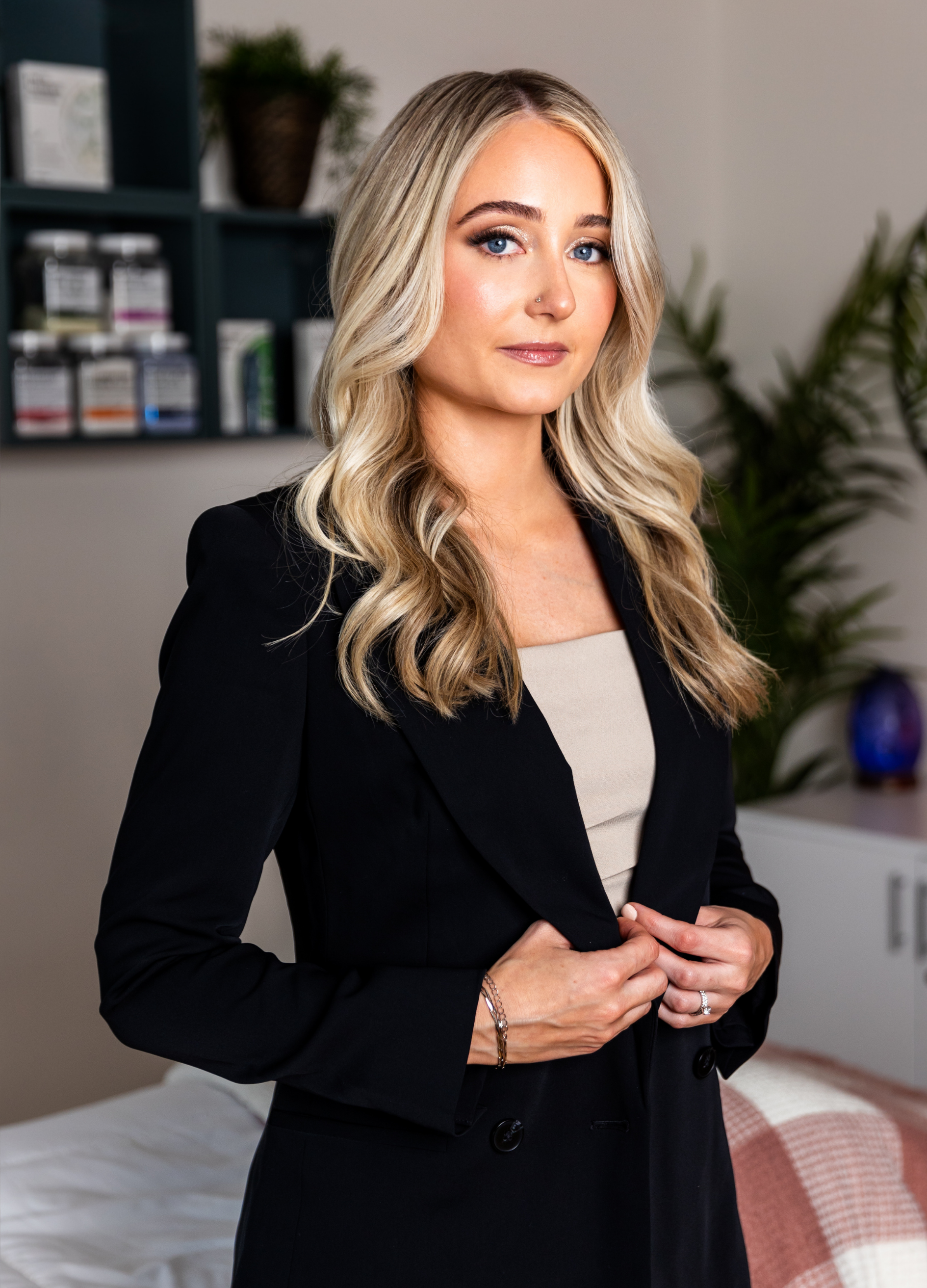 A confident woman with long, wavy blonde hair and blue eyes, dressed in a black blazer over a beige top, standing in a modern, well-lit room with plants and decorative items.