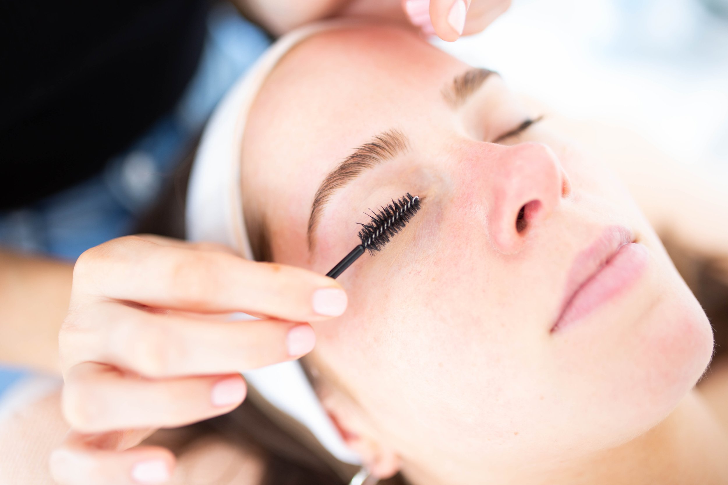 A woman receiving eyelash extension treatment, lying with eyes closed, while a technician applies mascara or lash extension with a tool.