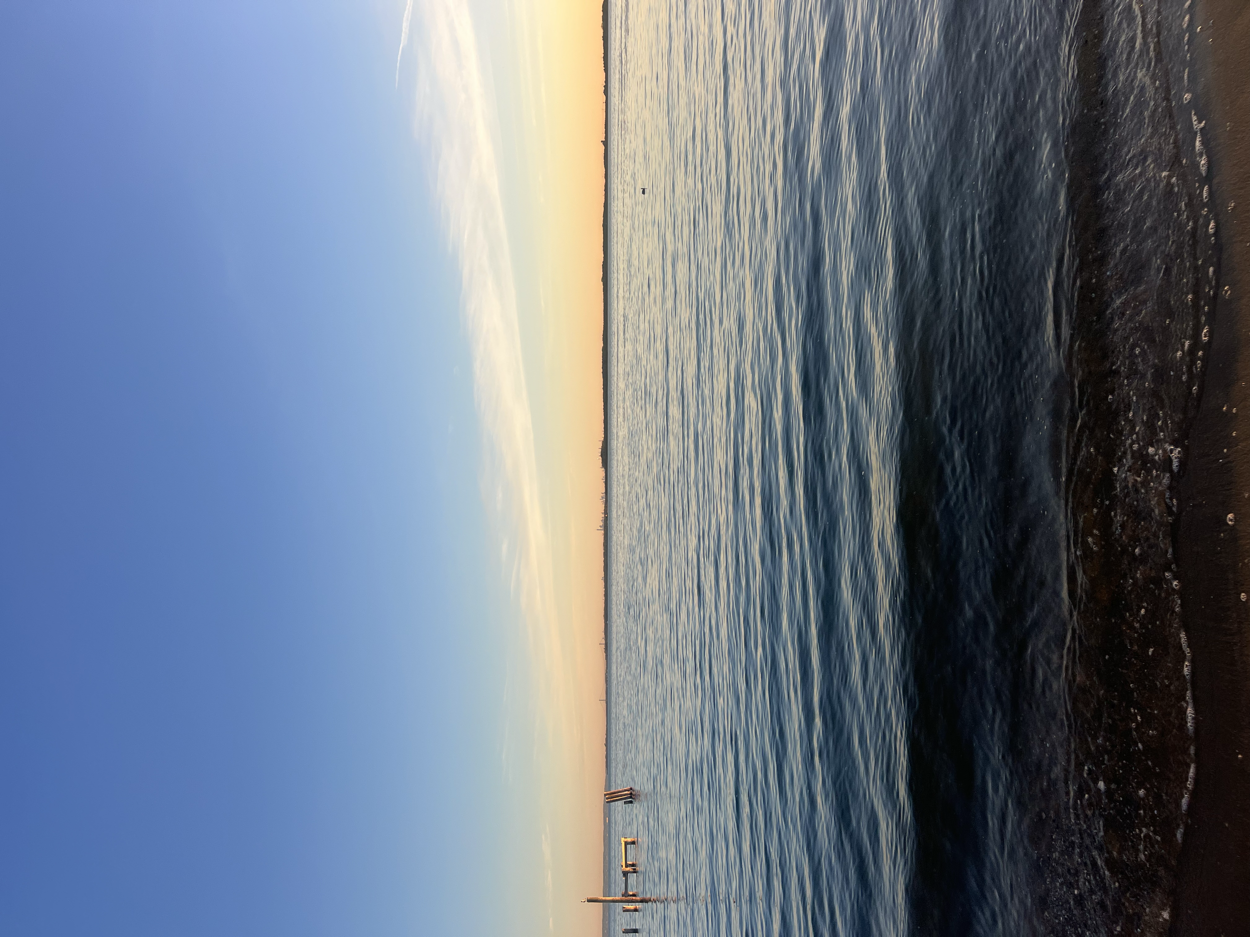 A calm body of water at sunset with a pastel-colored sky and sailboats in the distance. The shoreline is visible with rocks and wooden posts in the foreground.