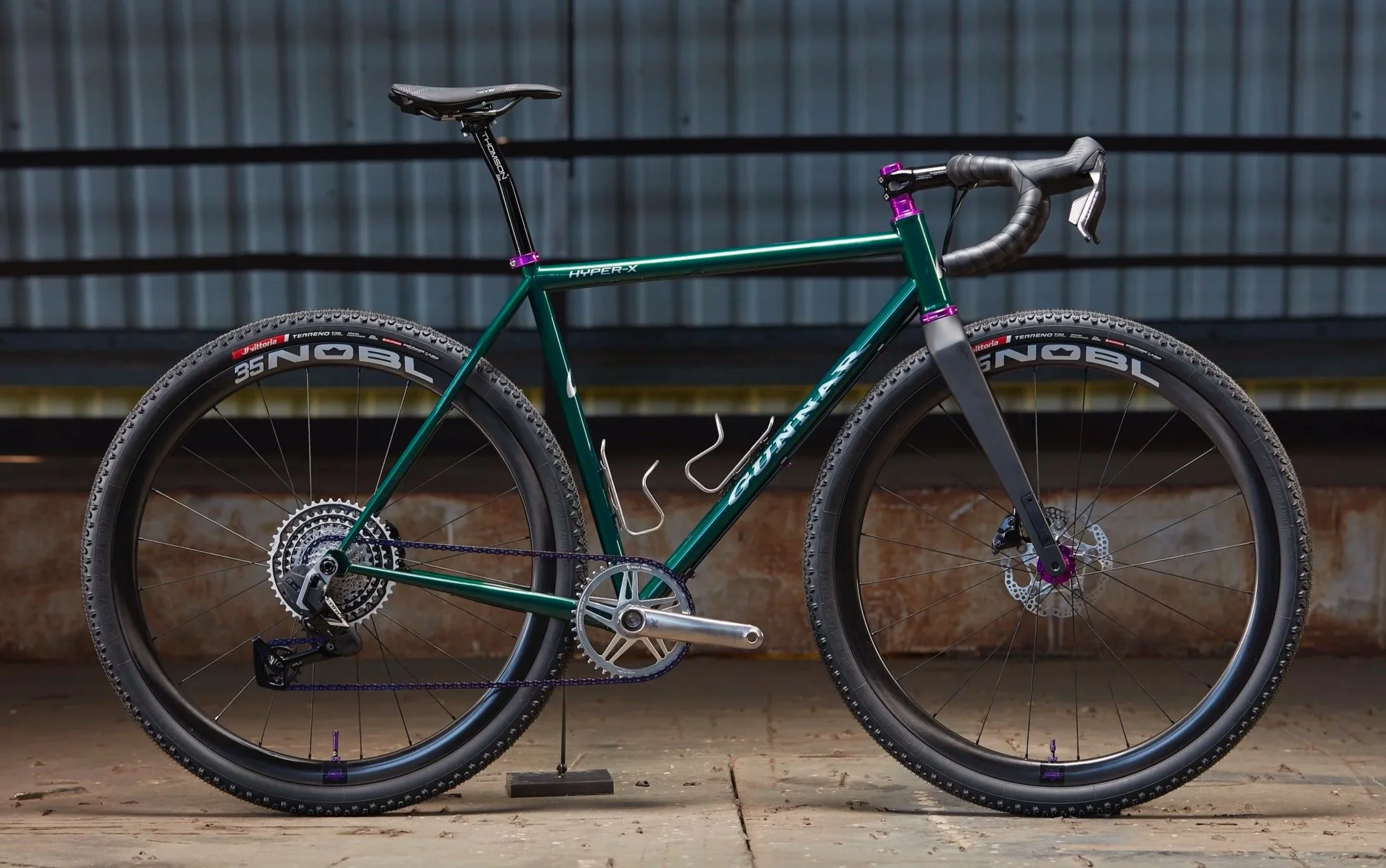 A green mountain bike with purple accents, drop handlebars, knobby tires, and disc brakes, is positioned indoors on a wooden floor with a metal wall in the background.