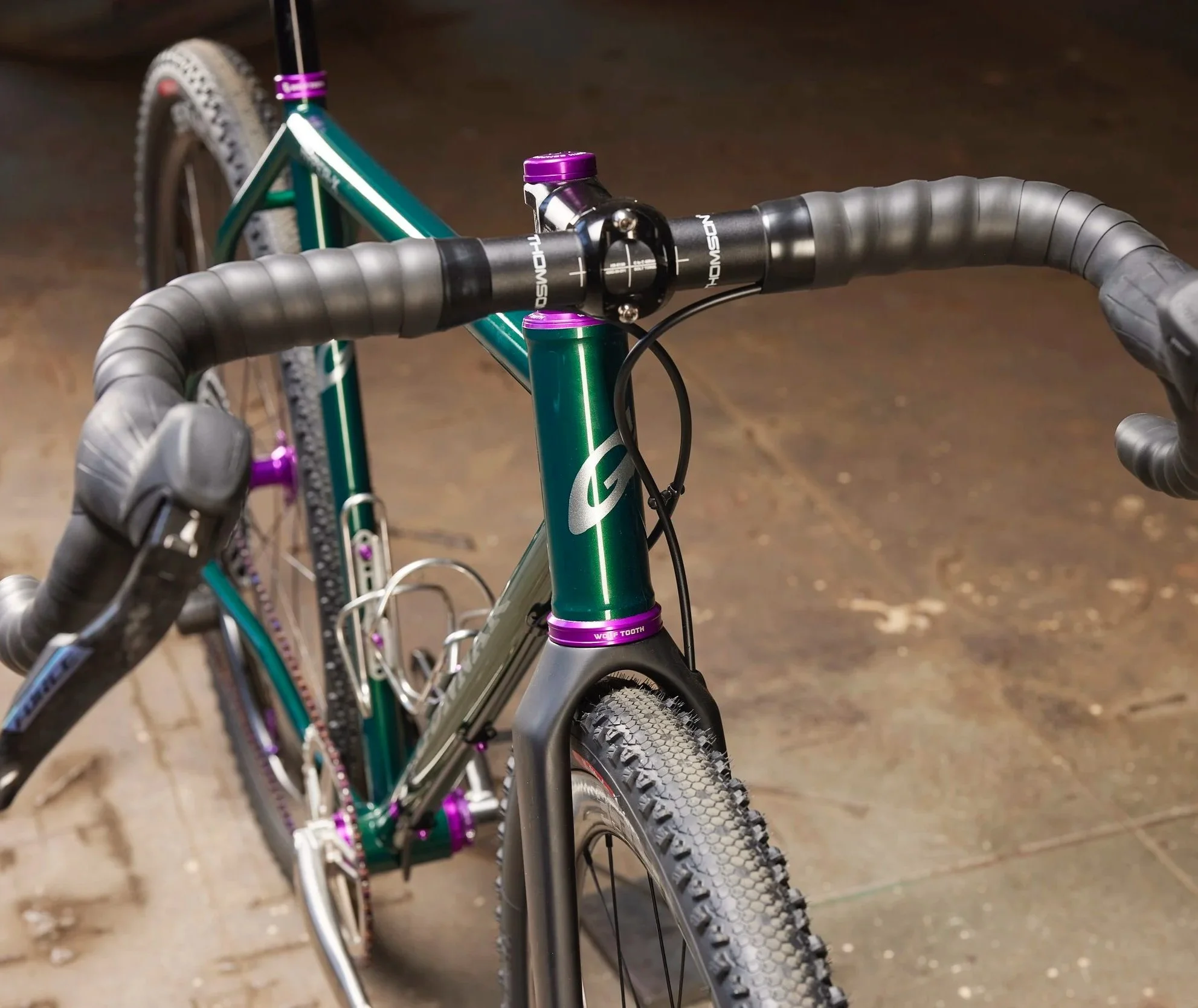 Close-up of a green mountain bike with a black handlebar and purple accents, focusing on the handlebar and front tire, with a dirt floor background.