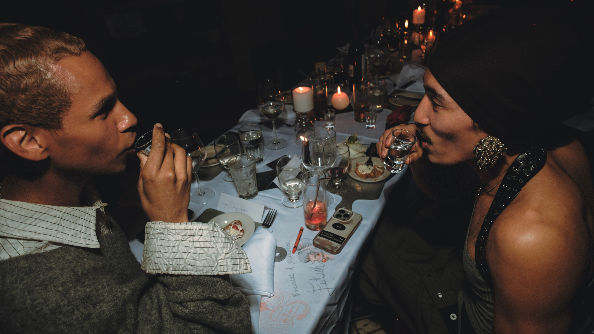 Two people sitting at a candlelit dinner table, drinking glasses in hand, engaging in conversation in a dimly lit setting.