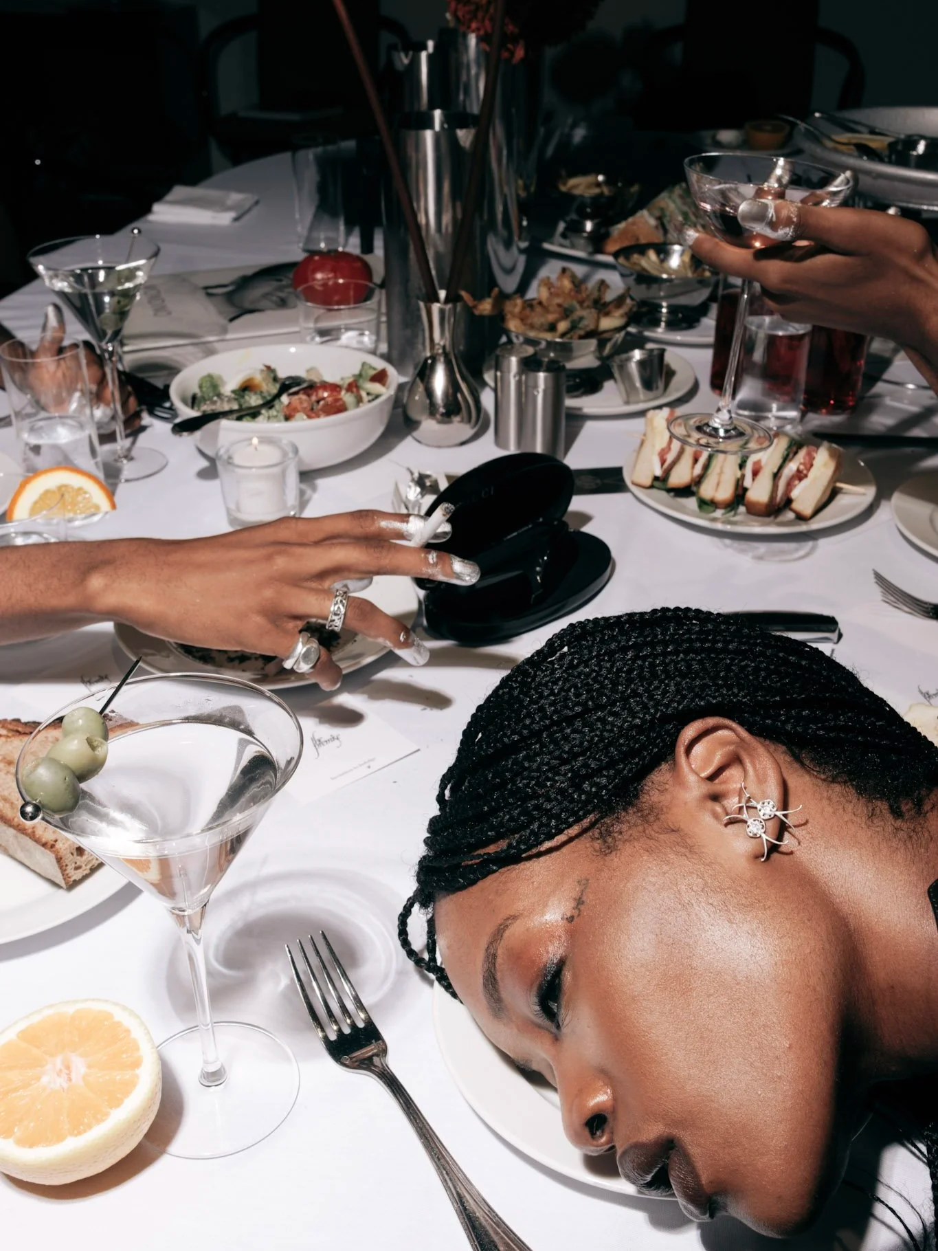 A woman with braids has her head on a white dinner plate at a dining table, with various dishes, drinks, and utensils, in a restaurant setting.