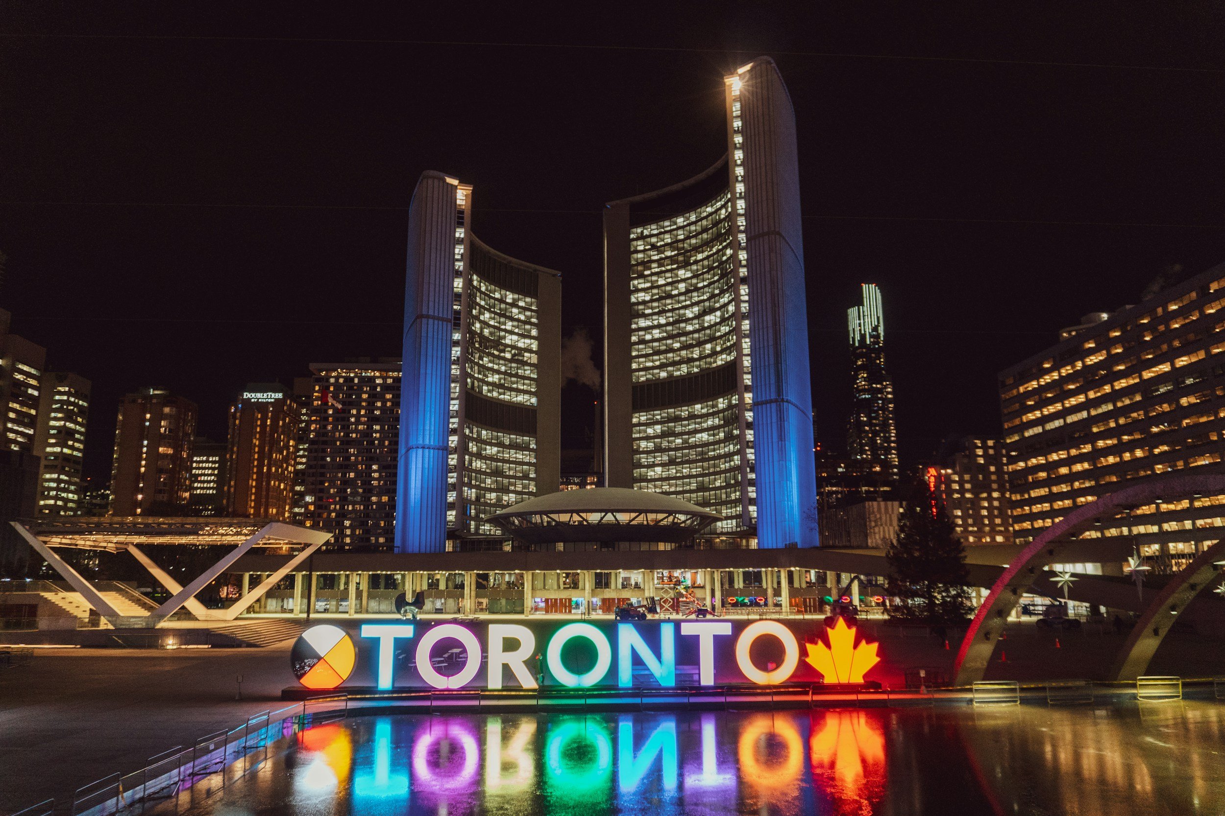 Night view of Toronto's skyline with the CS Touch tower illuminated, colorful 'TORONTO' sign reflected on a pond, and surrounding buildings lit up.