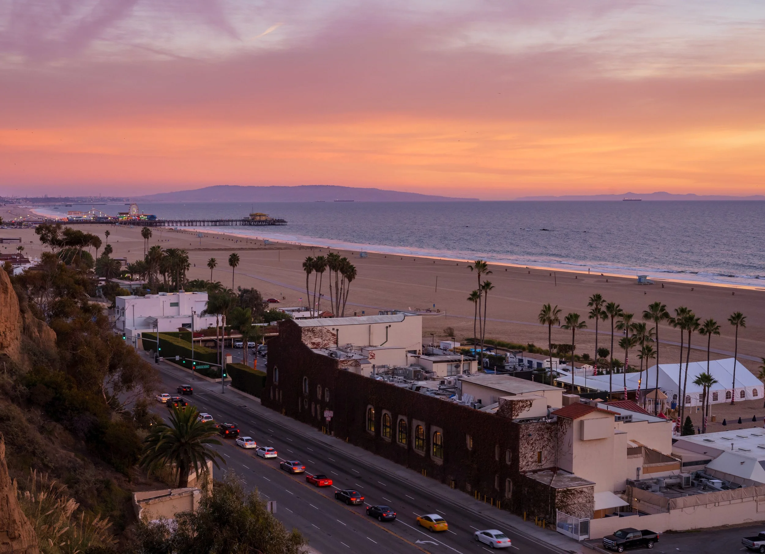 Sunset over a beach with palm trees and buildings, along a road with cars, and a pier extending into the ocean.