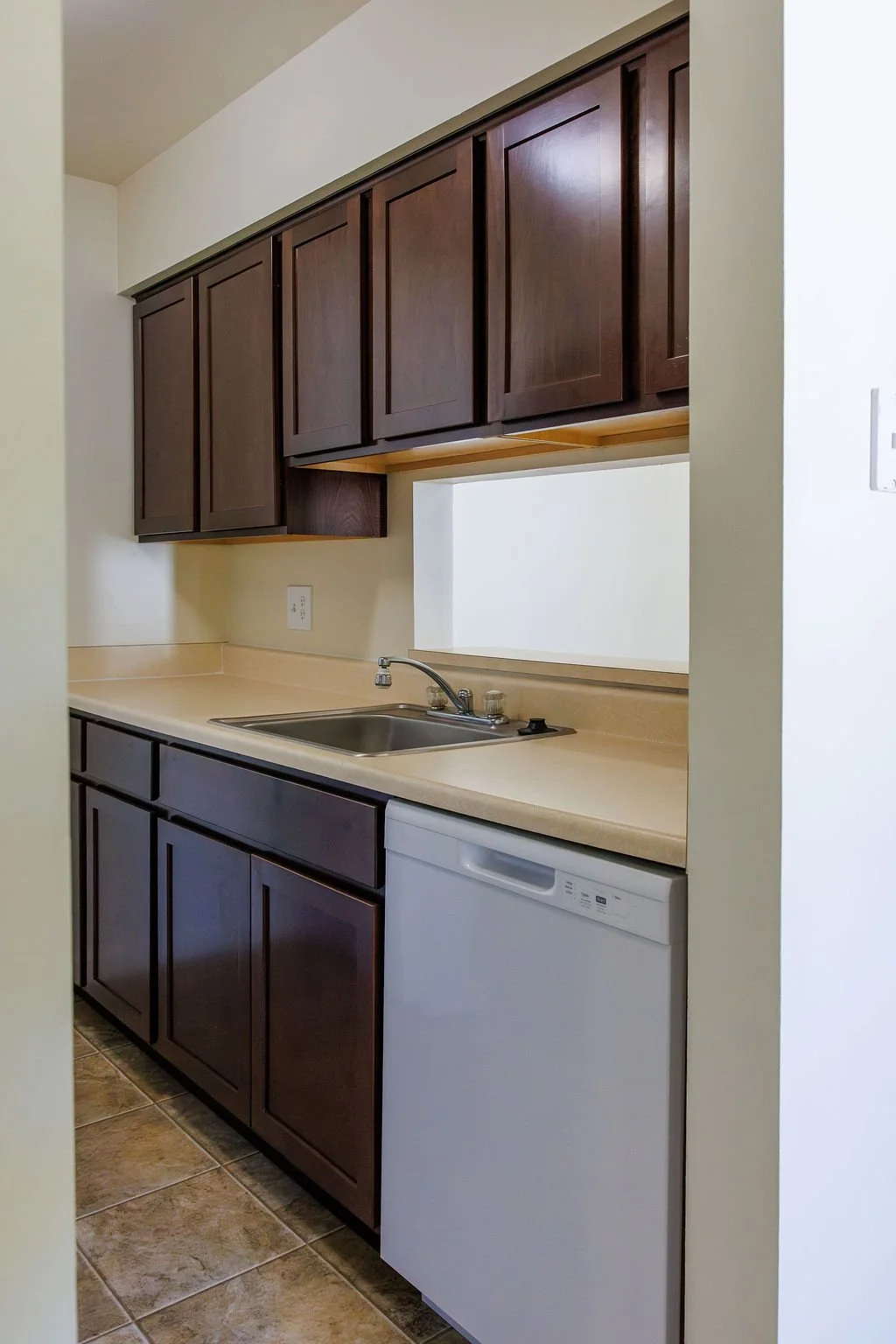 A small kitchen with dark wood cabinets, a beige countertop, a stainless steel sink with a chrome faucet, and a dishwasher.