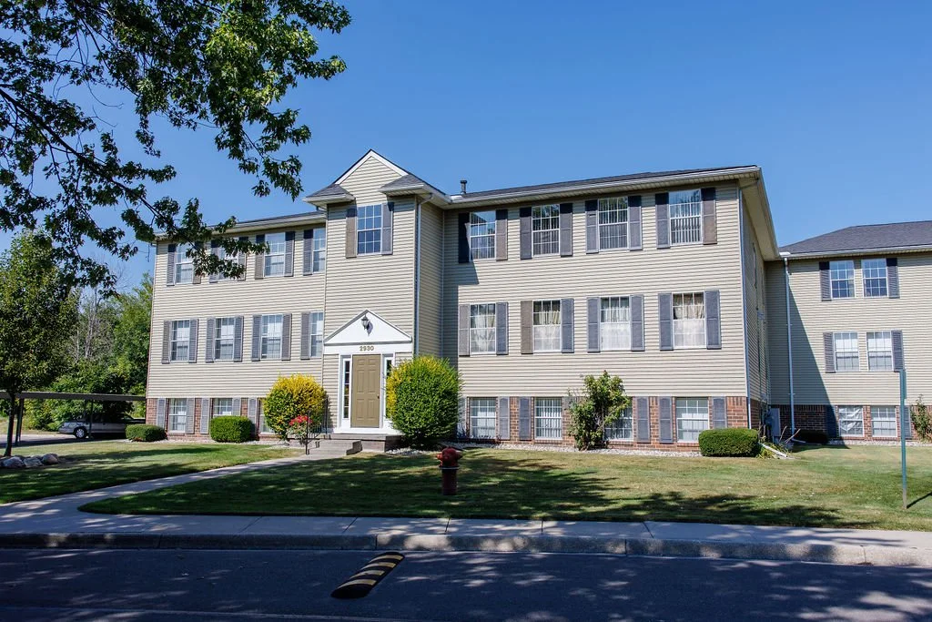 A multi-story residential building with beige siding, white trim, and black shutters, surrounded by a green lawn and trees under a blue sky.