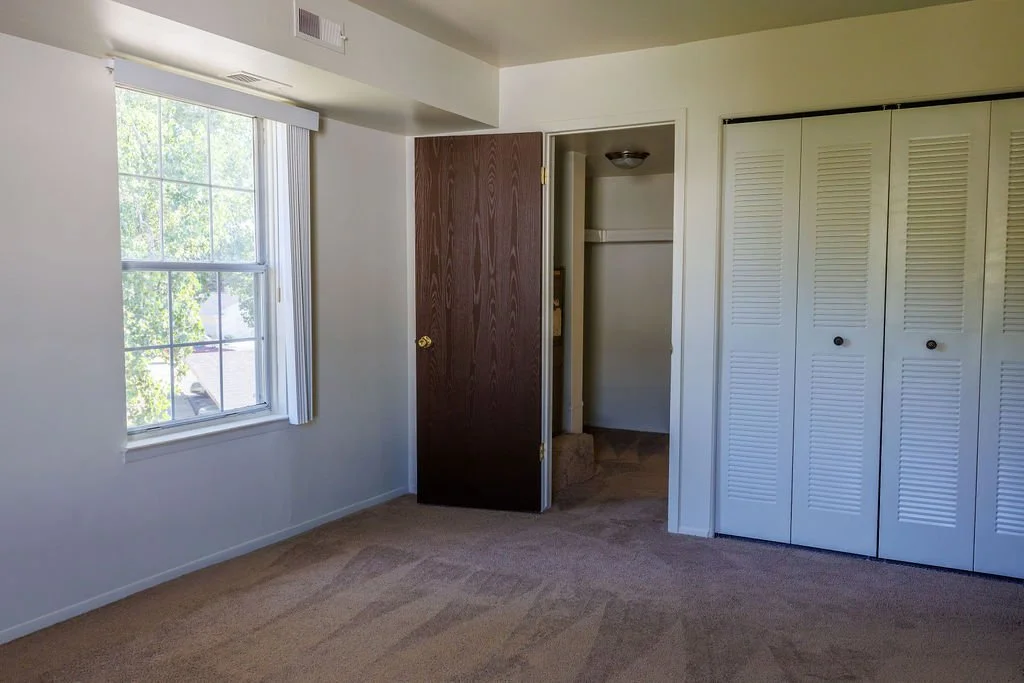 Empty bedroom with beige carpet, white walls, a window with white blinds, a dark brown door, and a white closet with bi-fold doors.