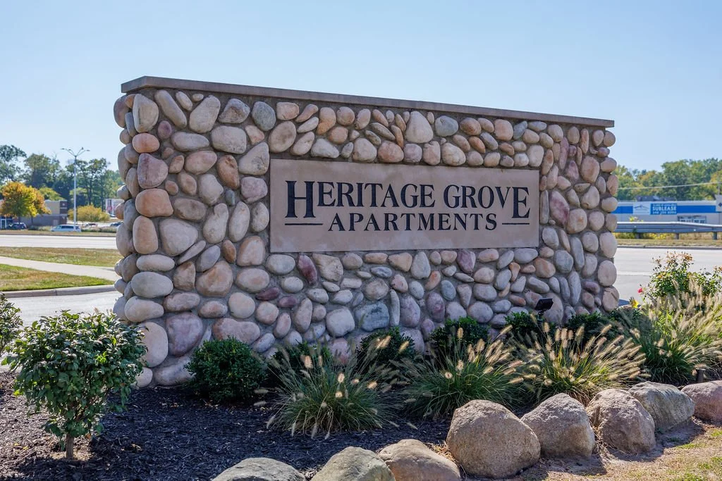 Sign for Heritage Grove Apartments made of stacked stones, with landscaped bushes and rocks in front.