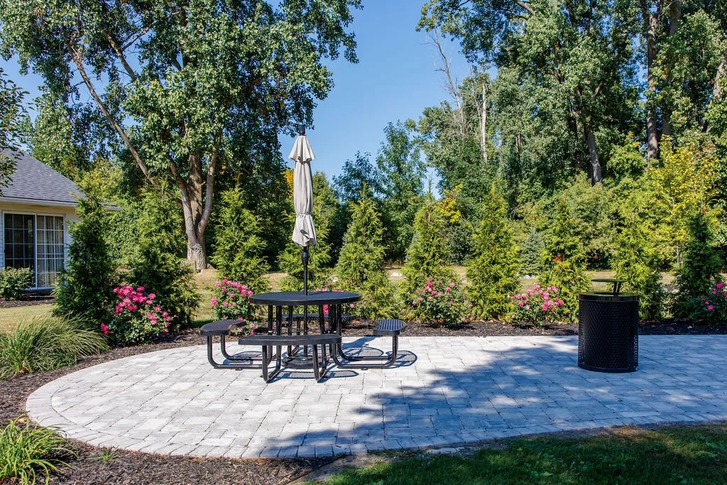 A backyard patio with a round metal table, attached bench seats, and a large closed umbrella, surrounded by lush green trees and flowering bushes, on a sunny day.