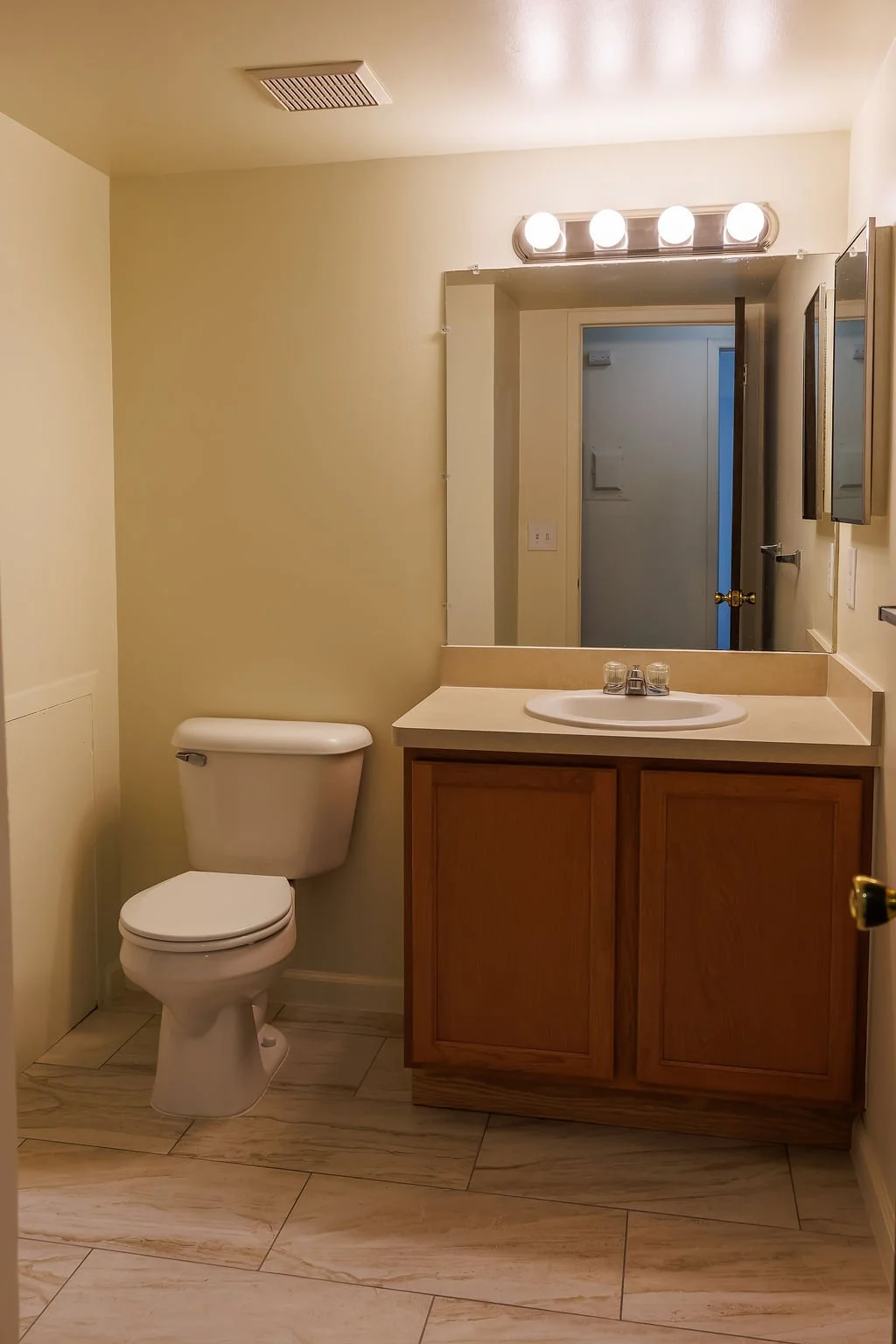 A small bathroom with a toilet, a wooden vanity with a sink, a large mirror, and a wall-mounted light fixture with four bulbs. The walls are painted a light cream color, and the floor has beige tiles with streaks.