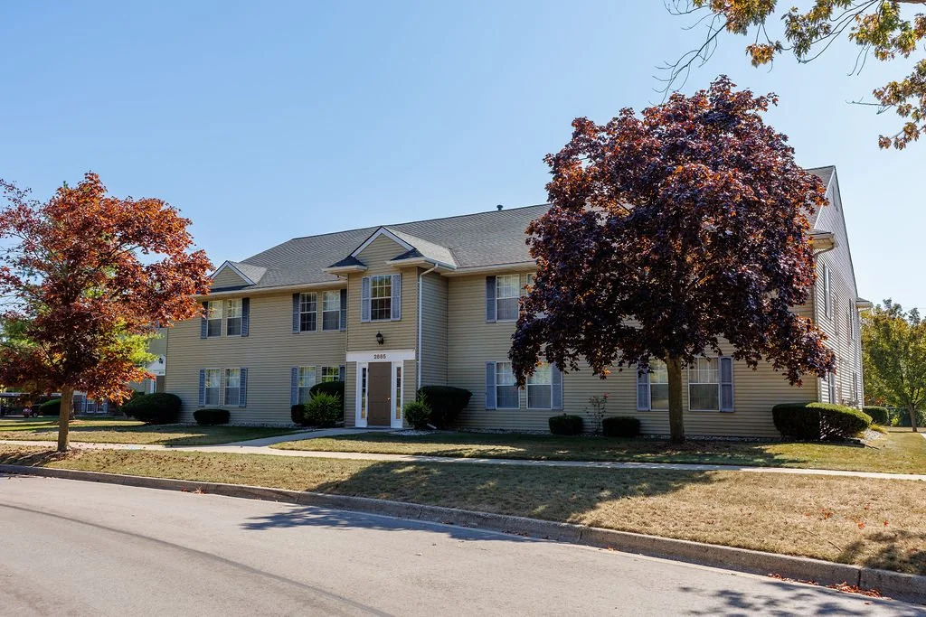 A two-story beige residential building with multiple windows and a gable roof, surrounded by trees with red and green leaves, on a street with a sidewalk and grassy lawns.