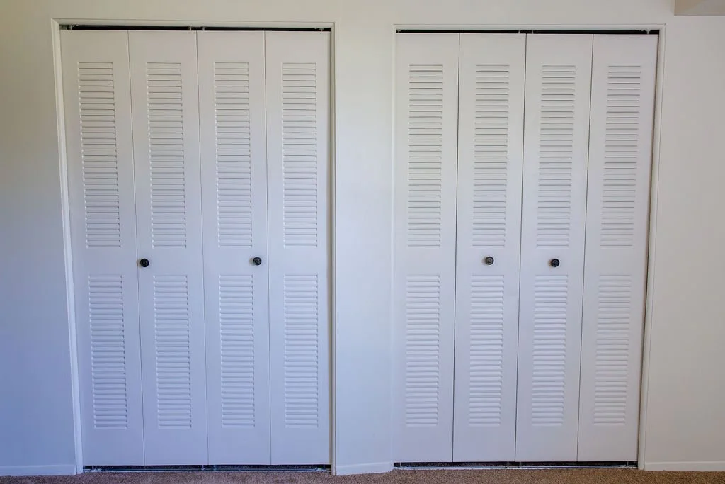 Two white closet doors with horizontal slats, each with a small round black knob, located in a room with beige carpet and white walls.