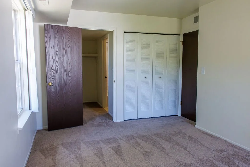 Empty bedroom with beige carpet, white walls, a window, a closed wooden door, and a closet with white bi-fold doors.